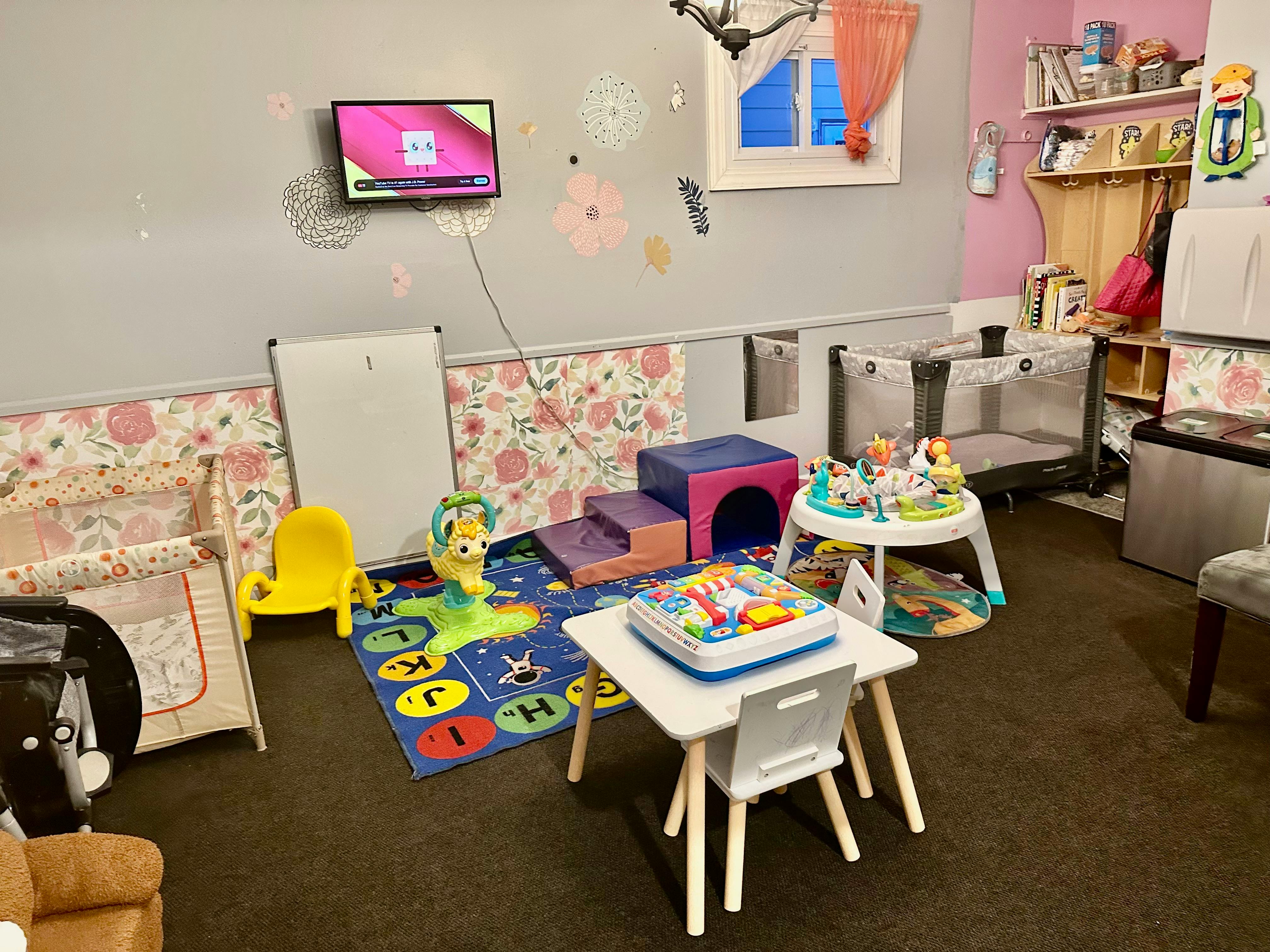 Playroom with cribs, playmats, small tables, and a wall-mounted television