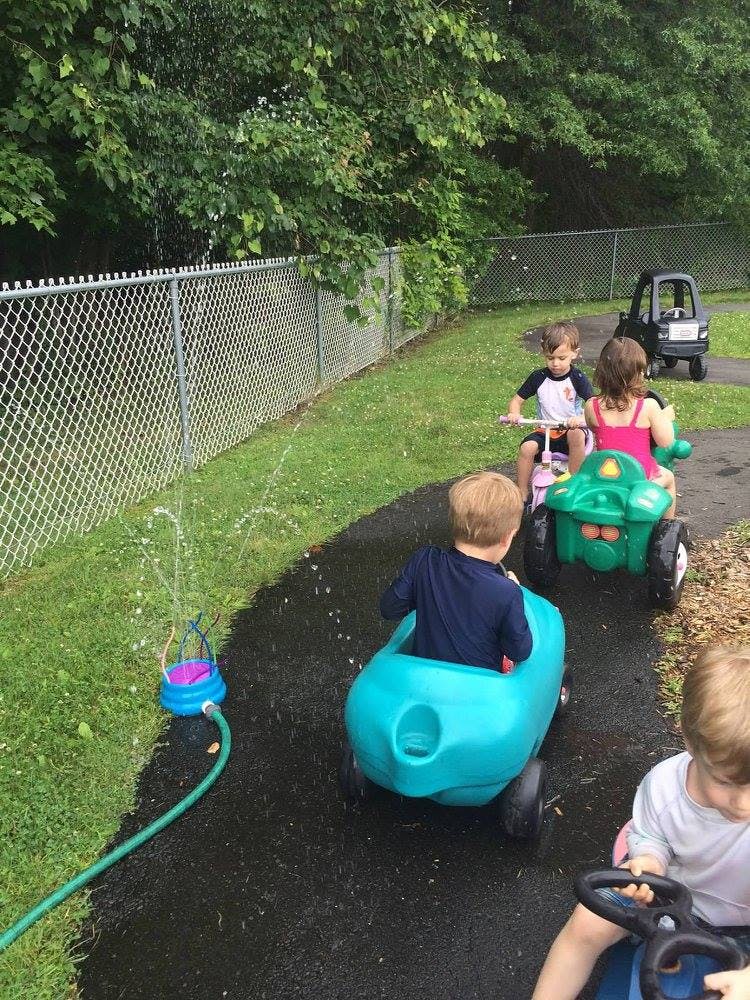 Children riding toy vehicles on a wet path with a sprinkler