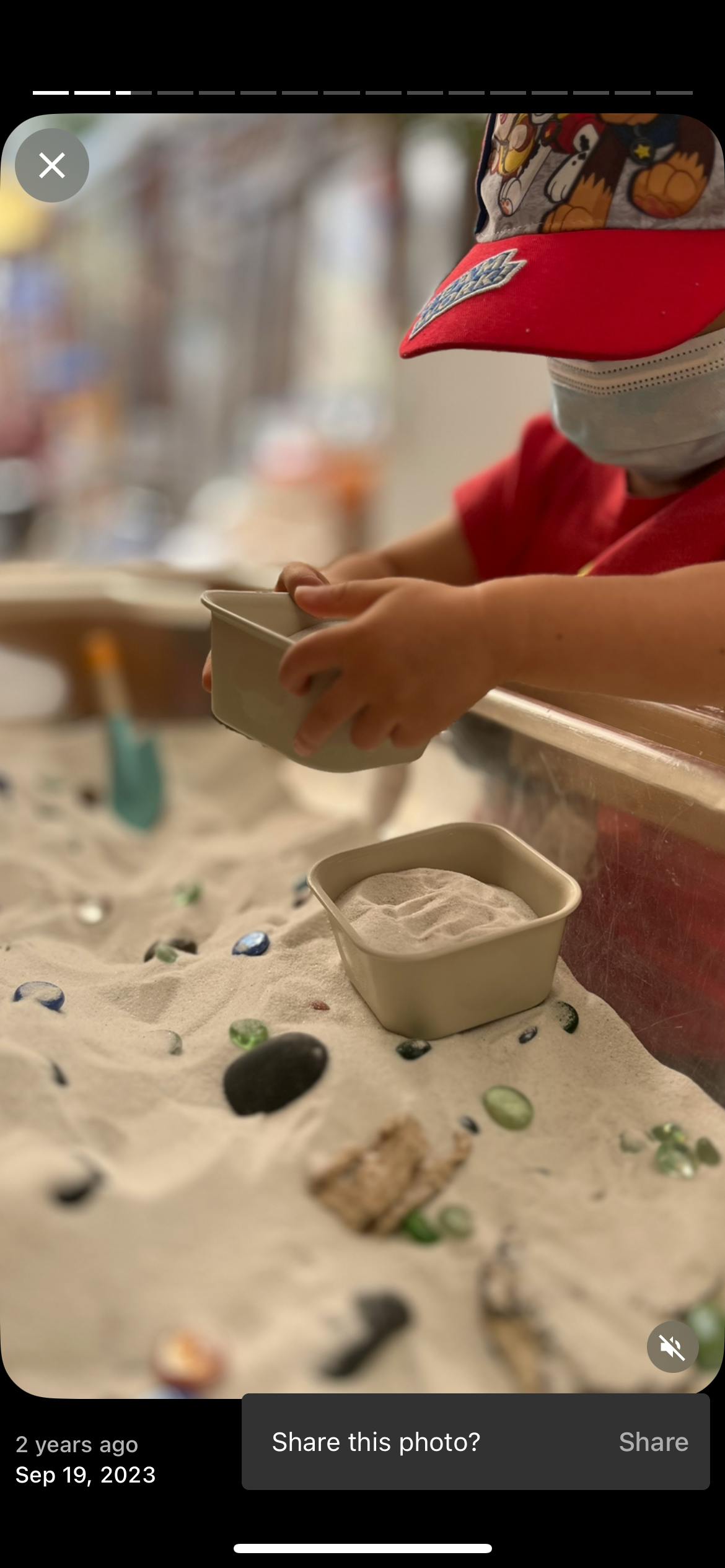 Child in a face mask and hat playing with sand and a small container