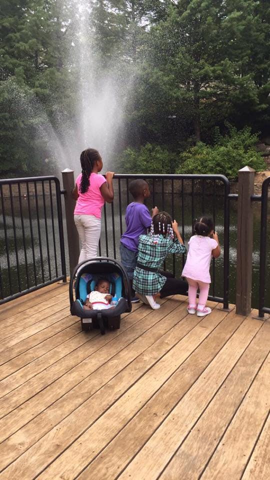 Children and a baby in a car seat on a wooden deck looking at a fountain