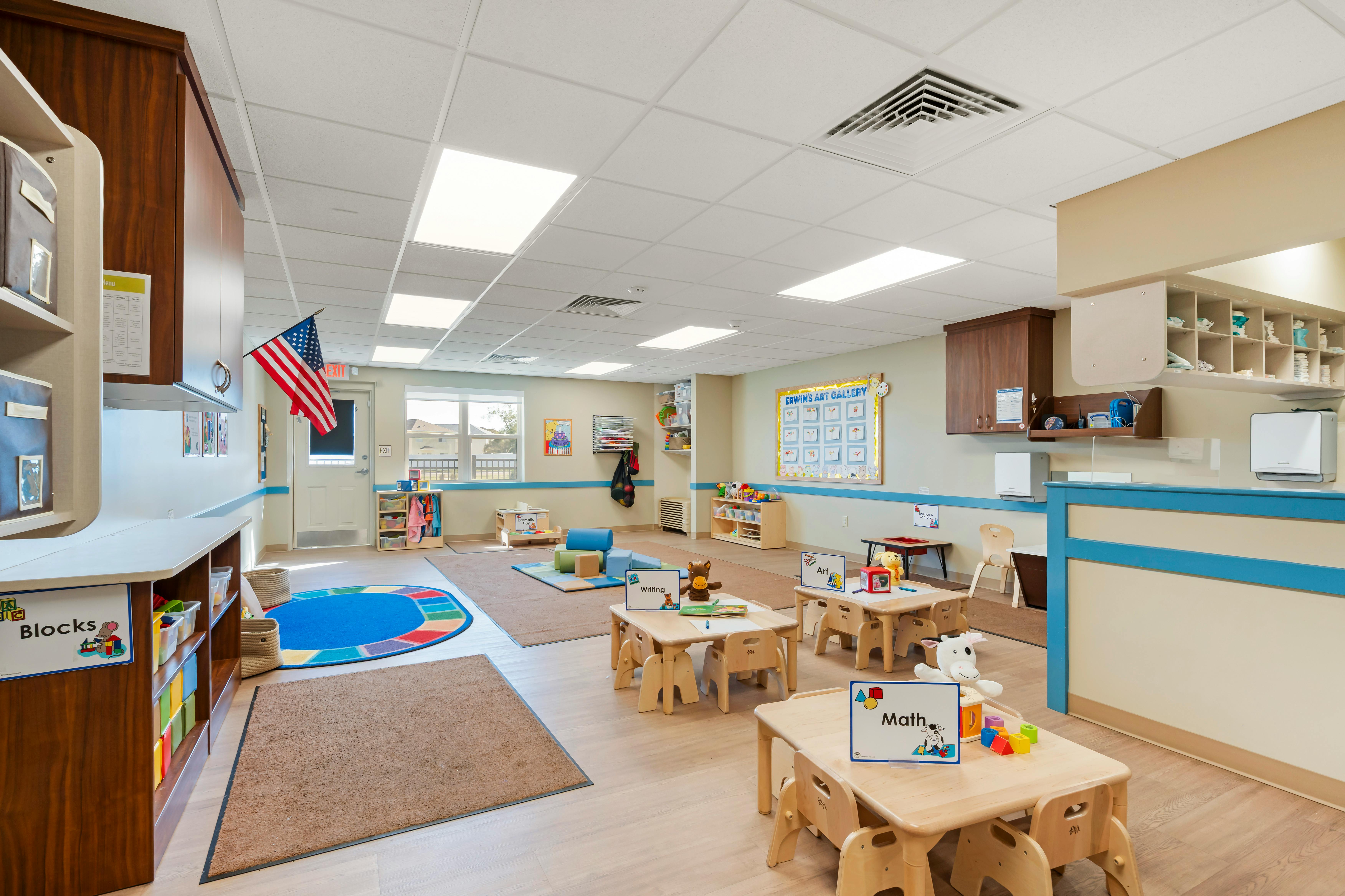 Empty classroom with small tables and chairs, play mats, and learning stations