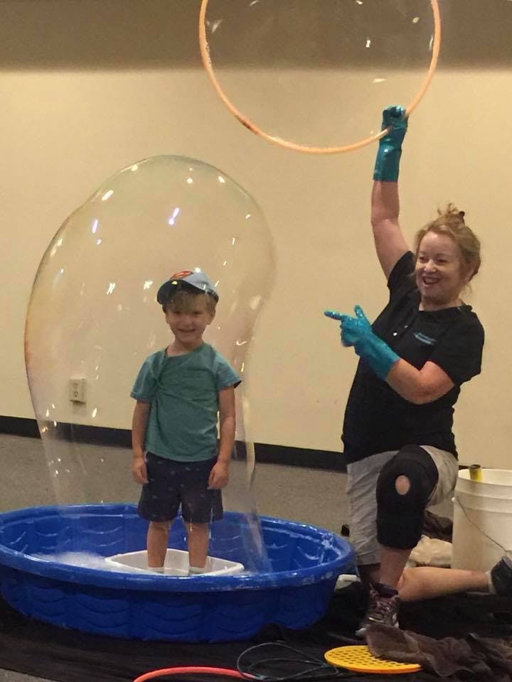 Person making a large bubble around a child standing in a tub