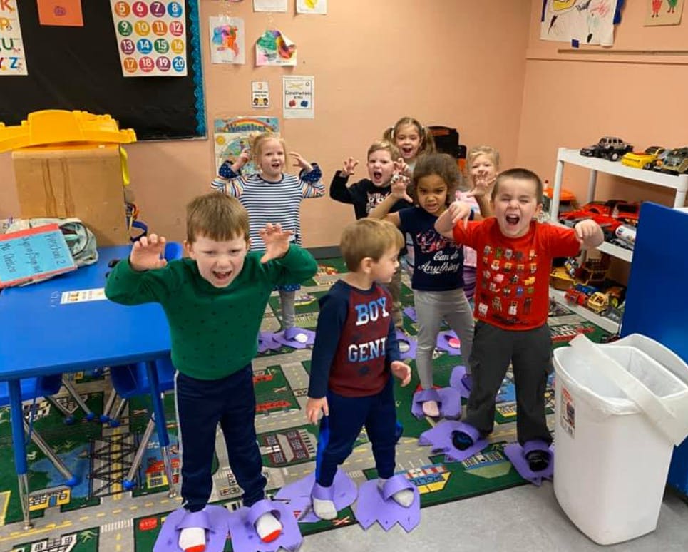 Group of children playing in a classroom, some wearing large purple shoe covers