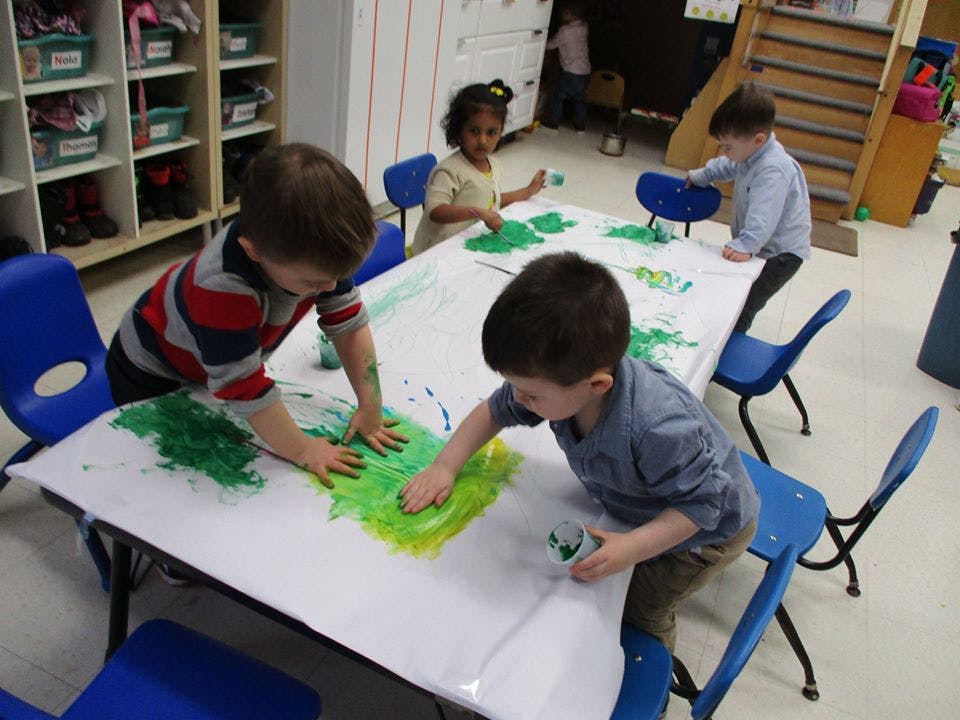 Children finger painting with green paint at a table in a classroom