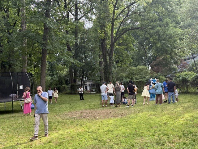 People gathered in a wooded backyard near a trampoline with a costumed character