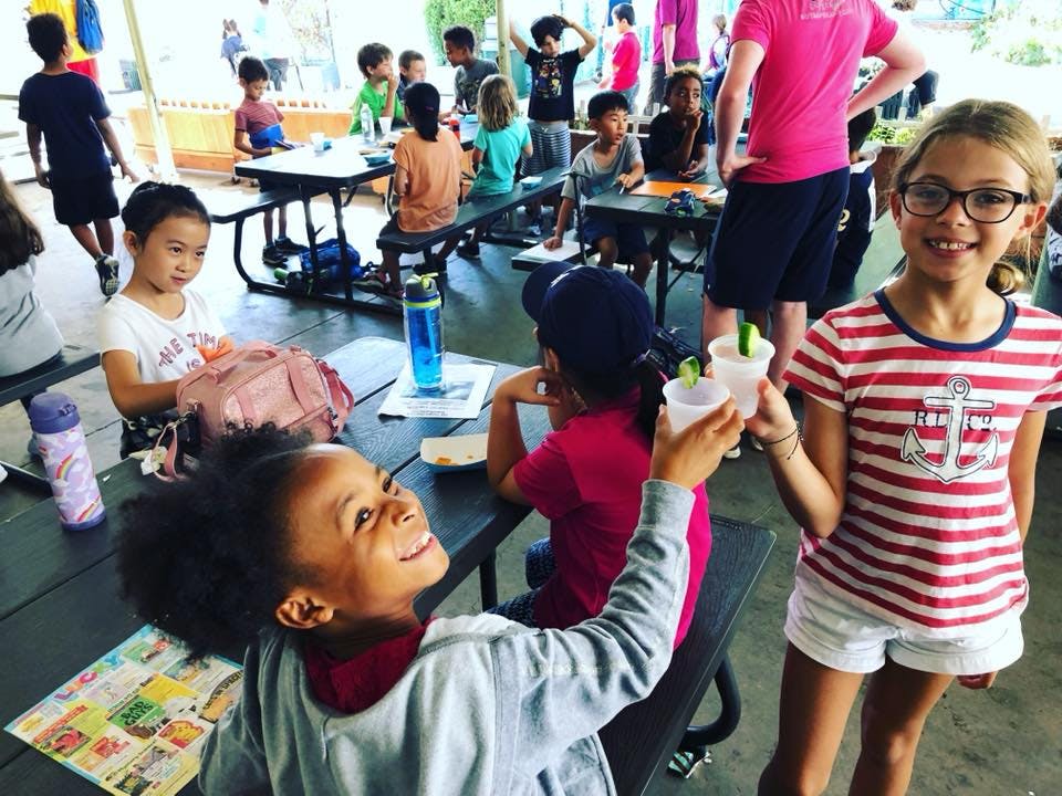 Two children clinking cups together, other children seated at tables in background