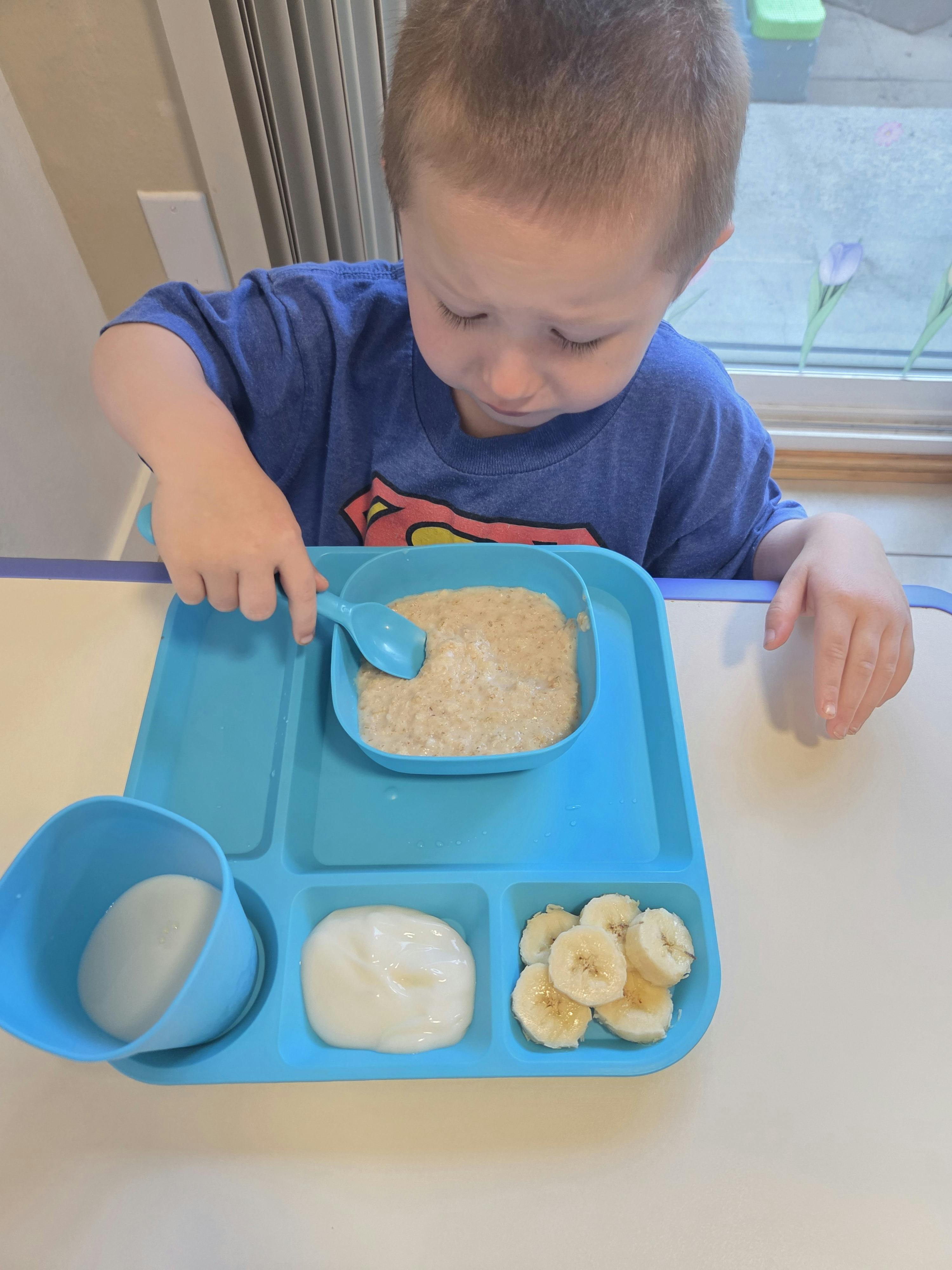 Child eating oatmeal with a spoon from a tray with yogurt and banana slices