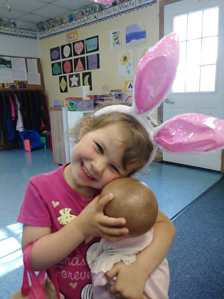 Child wearing bunny ears holding a doll in a classroom
