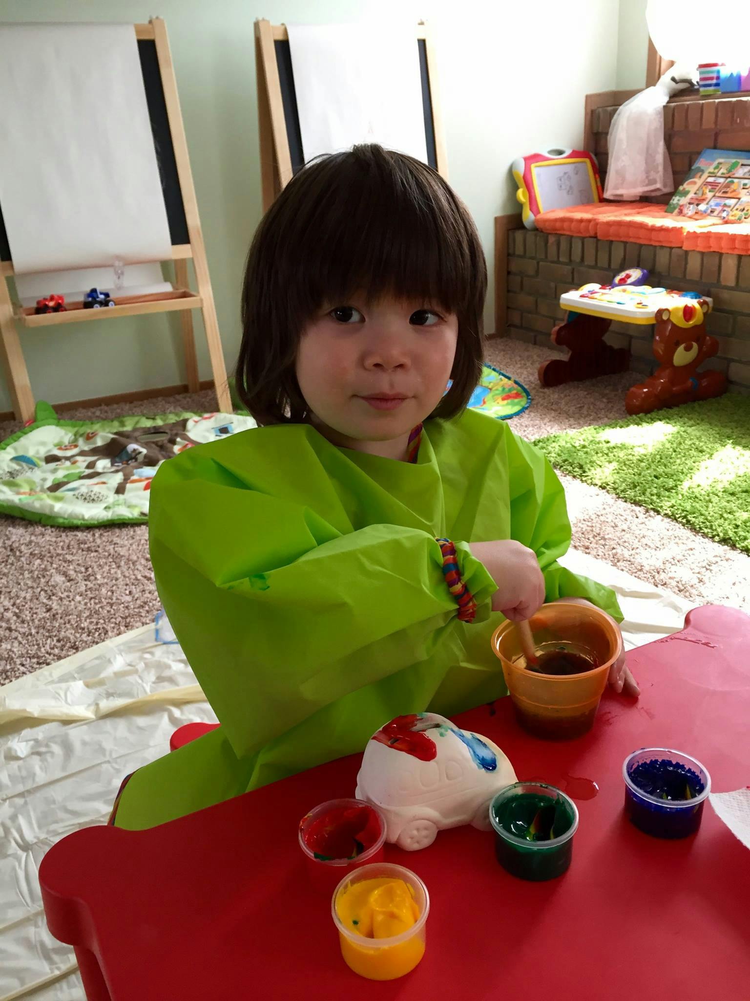 Child in a green smock painting a toy car at a red table