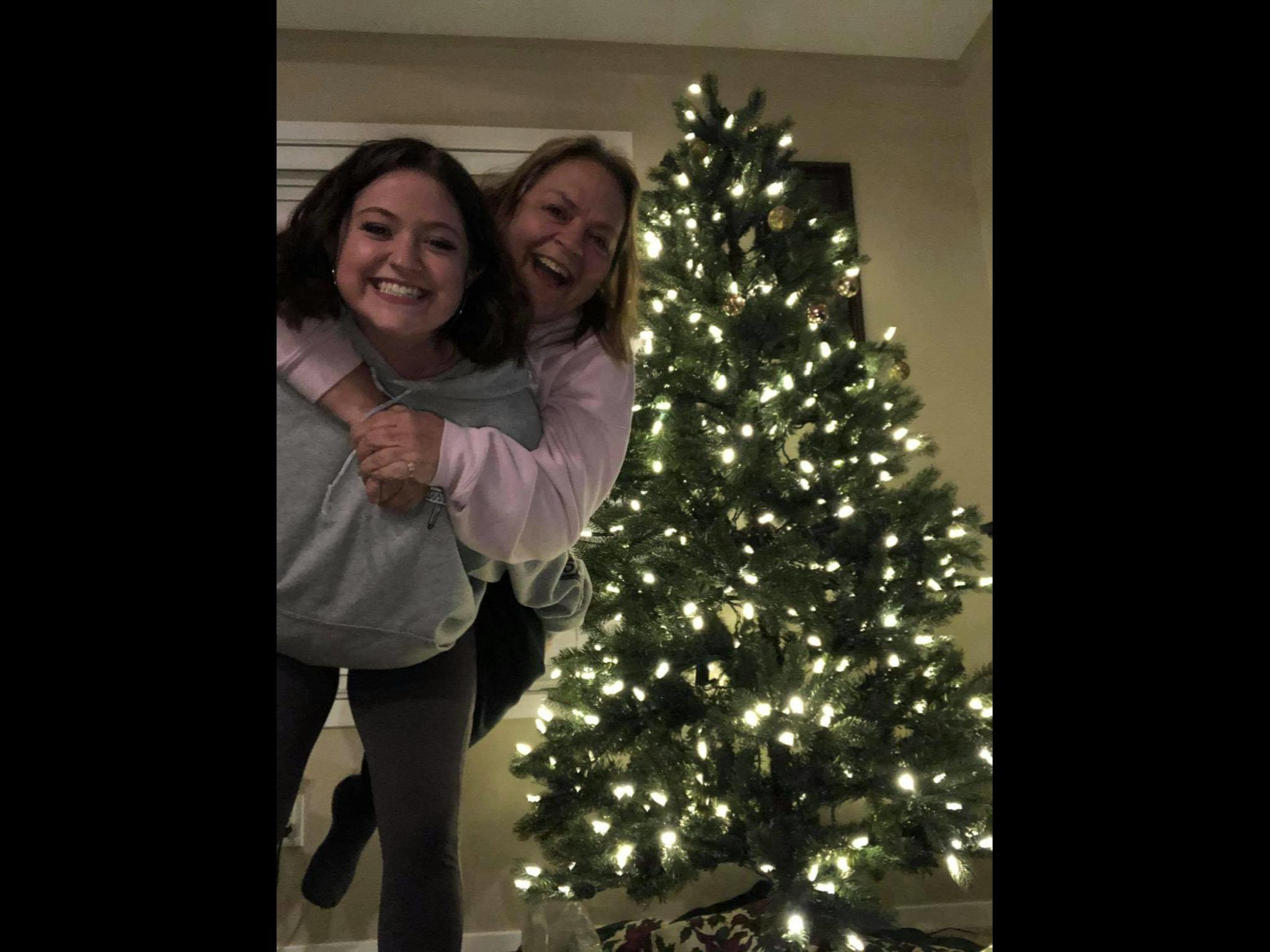 Two people piggybacking in front of a lit Christmas tree