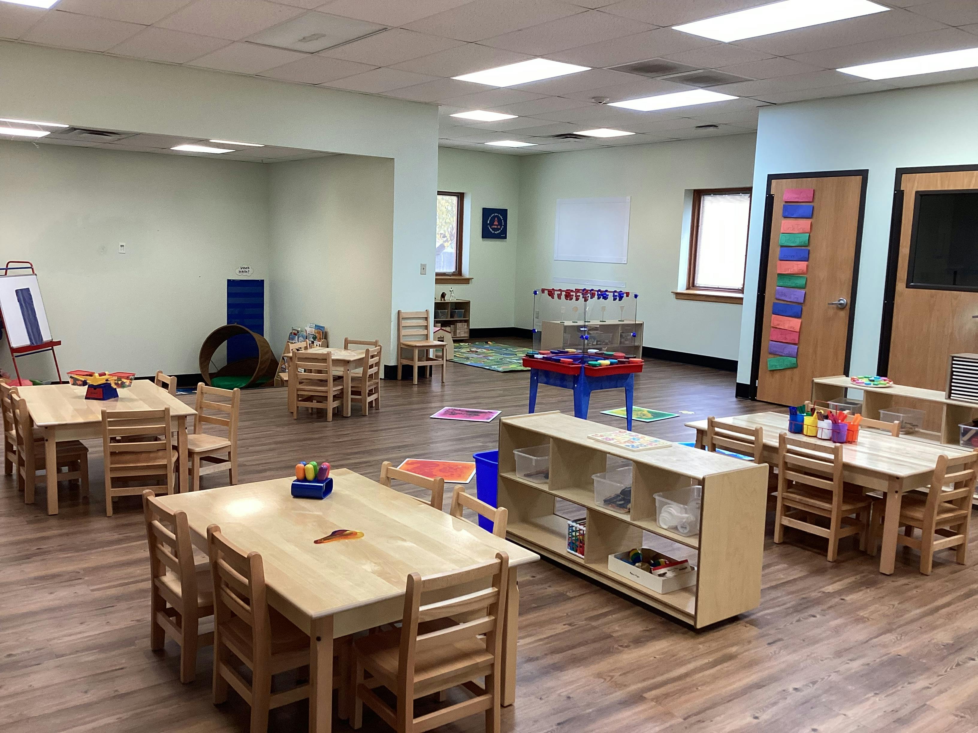 An empty classroom with light wood tables and chairs, shelves, and a foosball table