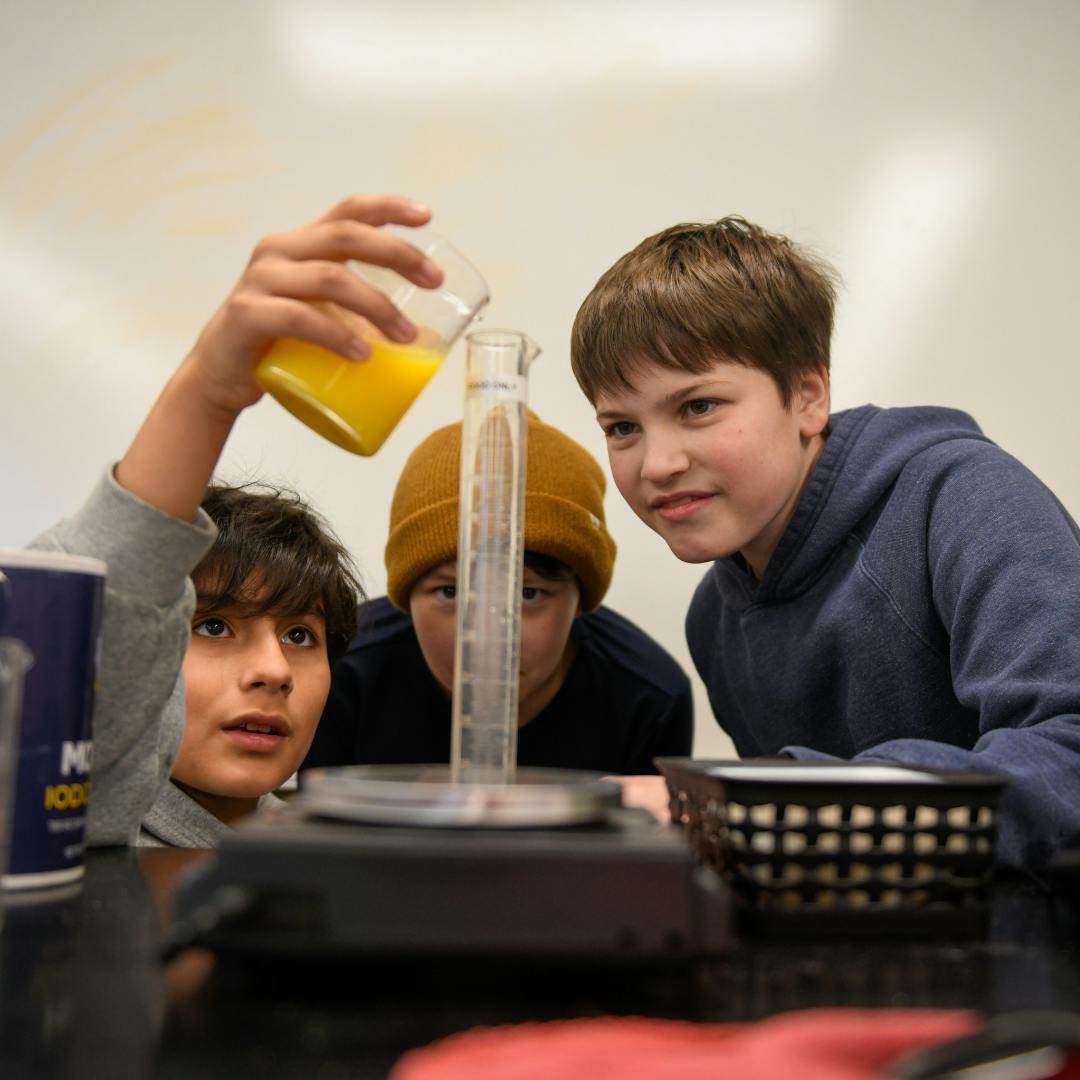 Three children in a classroom pouring liquid into a measuring cylinder