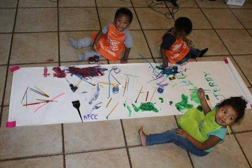 Three children sitting on a tiled floor, painting on a large sheet of paper