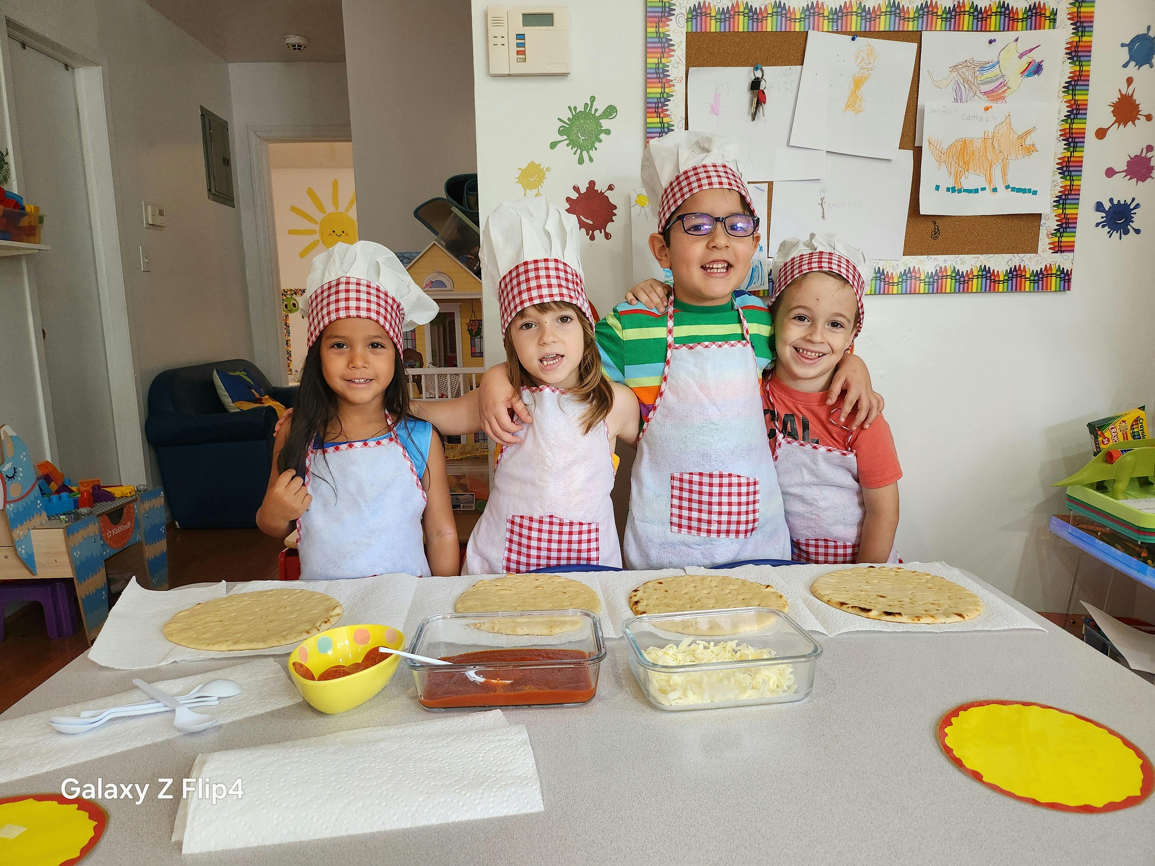 Four children in chef hats and aprons standing behind a table with pizza crusts