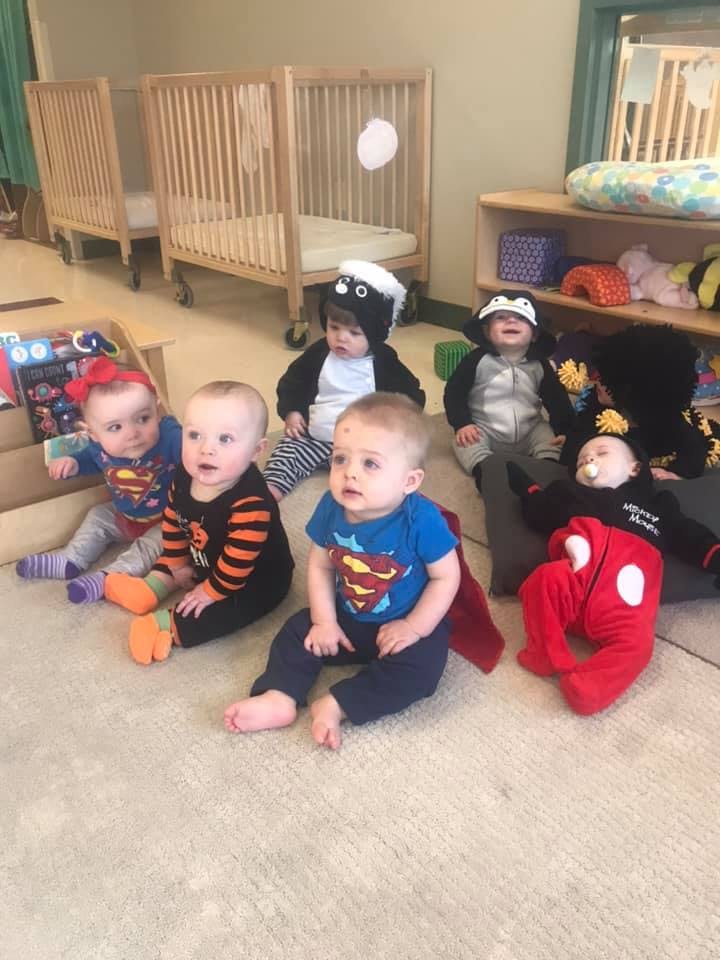 Multiple babies in various costumes sitting on a rug in a room with cribs