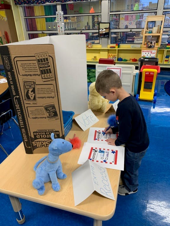 Child writing on a paper with "VOTE" on it in a classroom