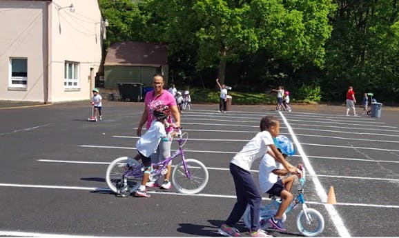 Adults helping children ride bicycles in a paved area