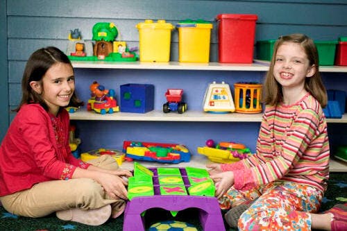 Two children playing with toys in a room with a shelf of toys in the background
