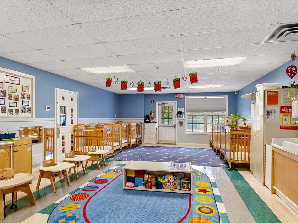 Childcare room with multiple cribs, small tables, and a colorful rug