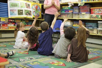 Children sitting on a rug with hands raised in a classroom