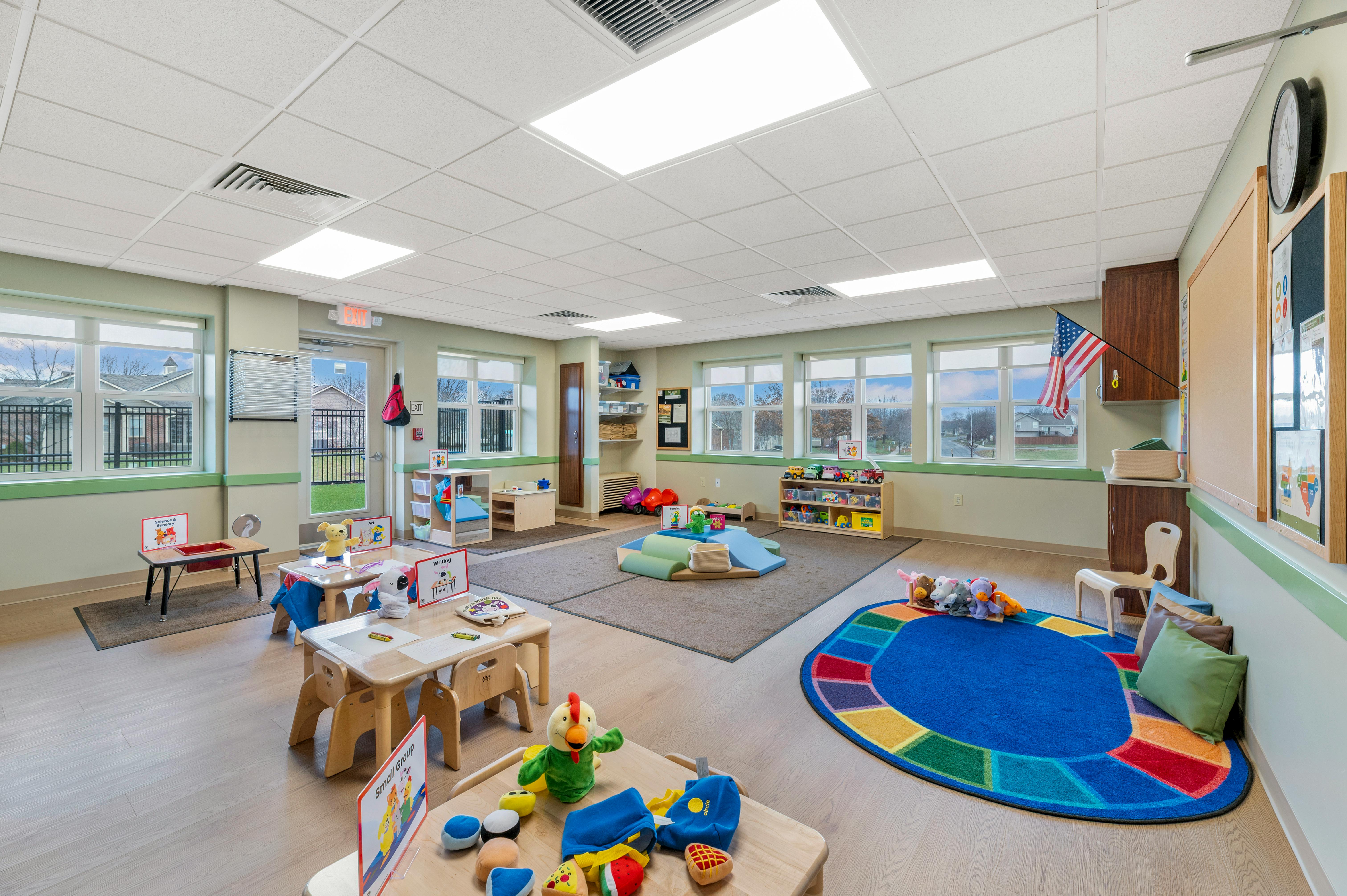 An empty classroom with small tables, chairs, colorful rugs, and play areas