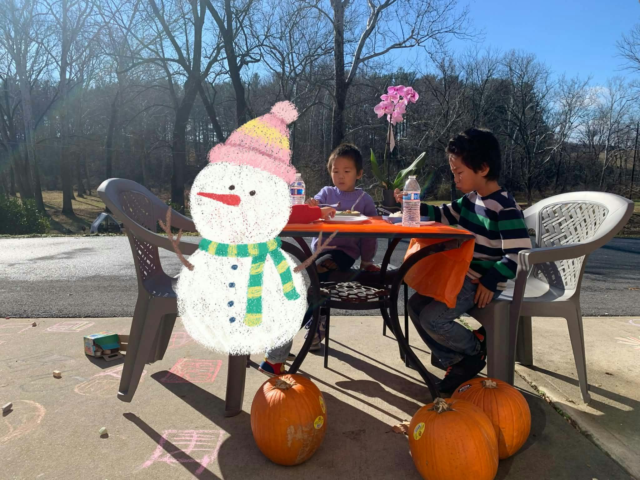Two children sitting at a table outdoors with a snowman cutout and pumpkins