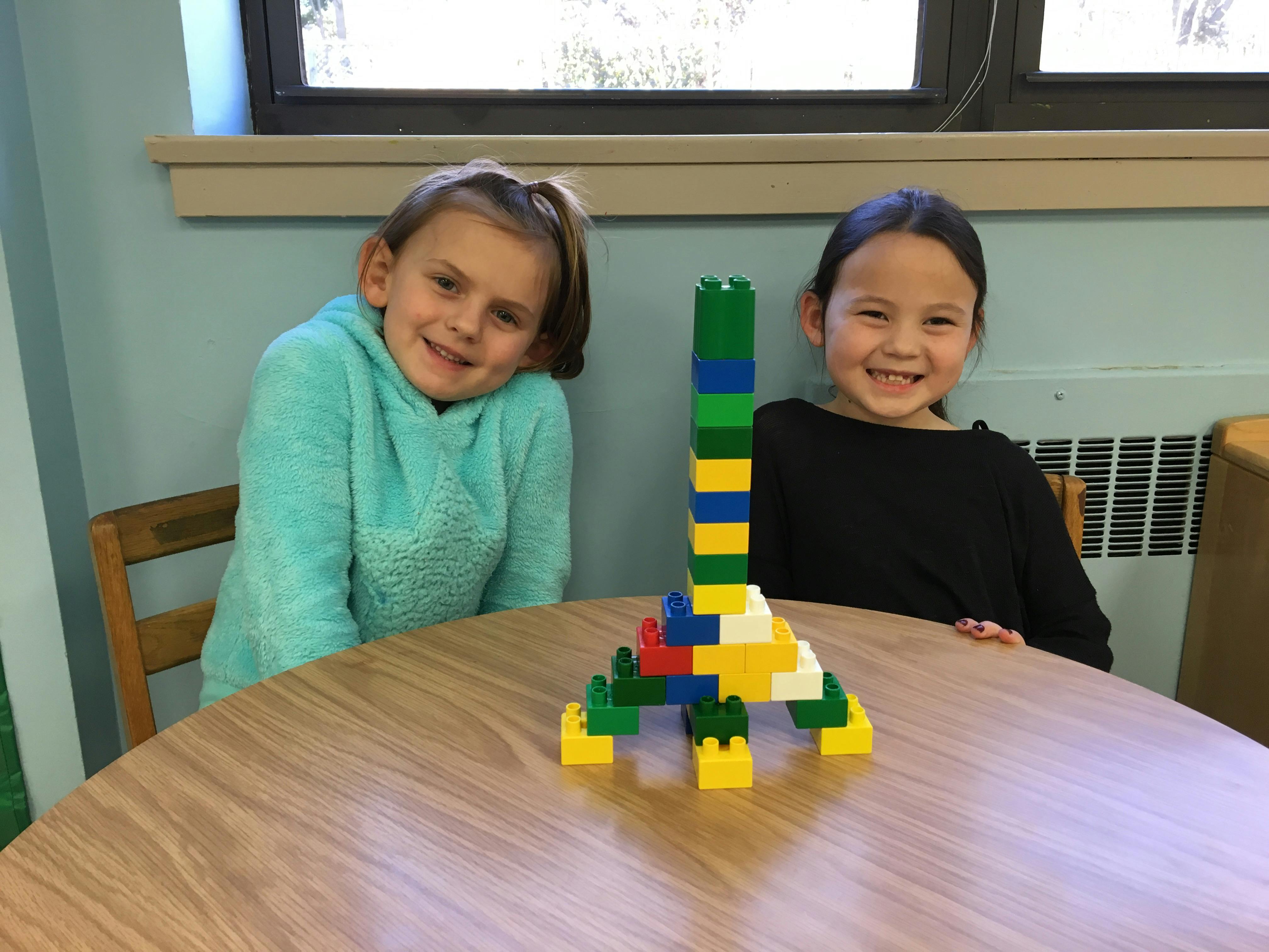Two children sitting at a table with a tall structure made of building blocks