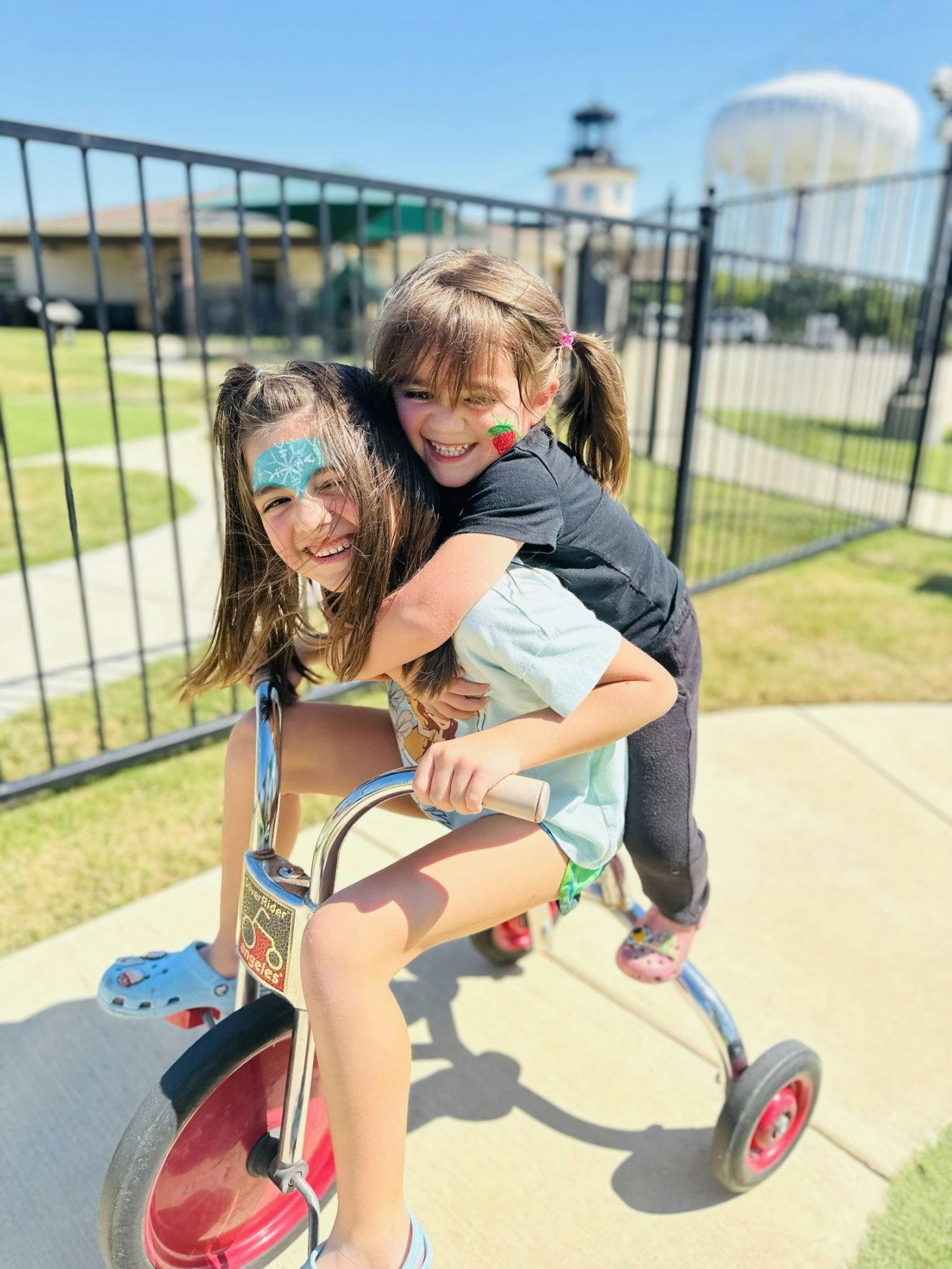 Two children with face paint riding a tricycle together outside