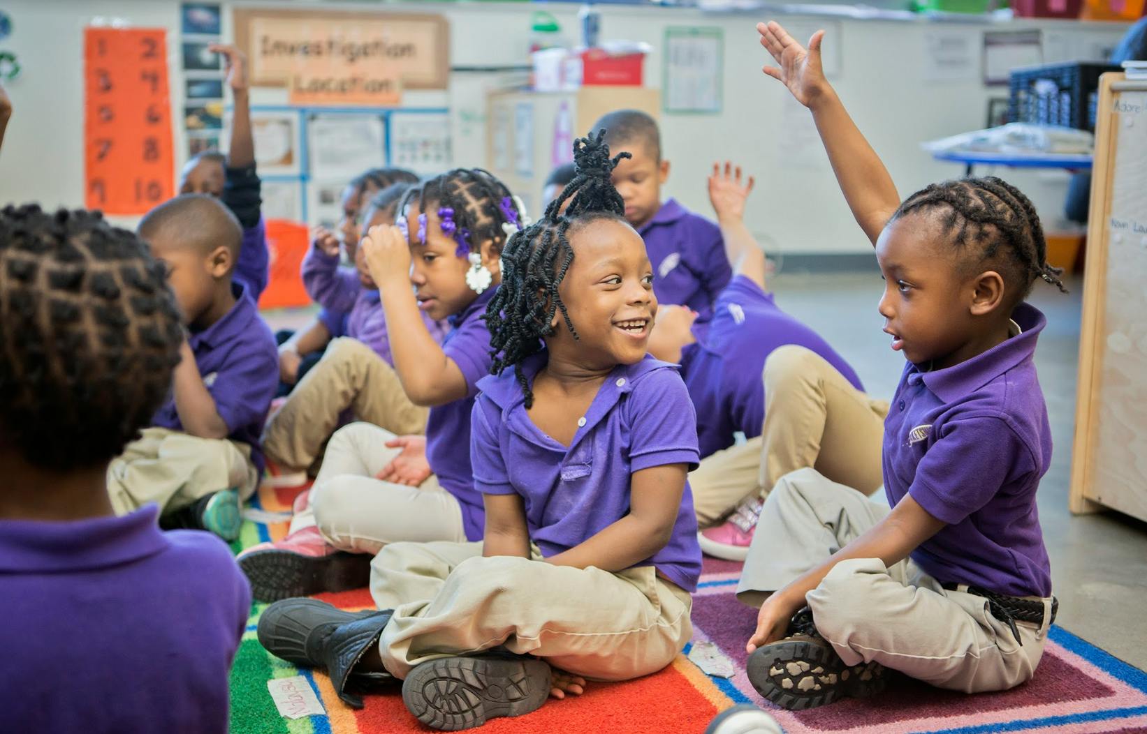 Children sitting on a rug in a classroom, some with hands raised