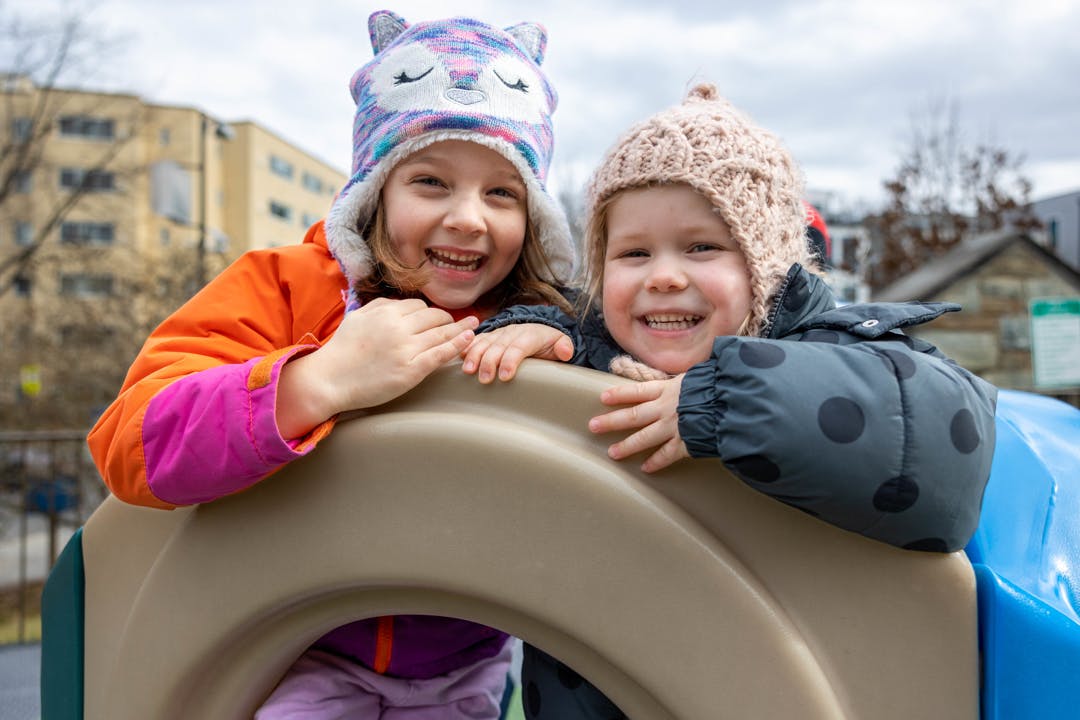 Two children in winter hats and coats peeking over playground equipment