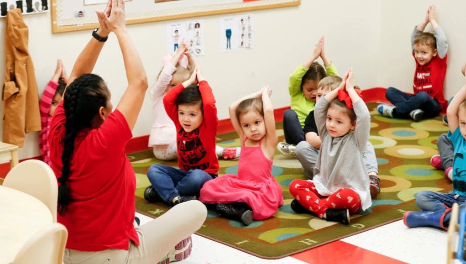 Person and children sitting on floor with arms raised in a classroom