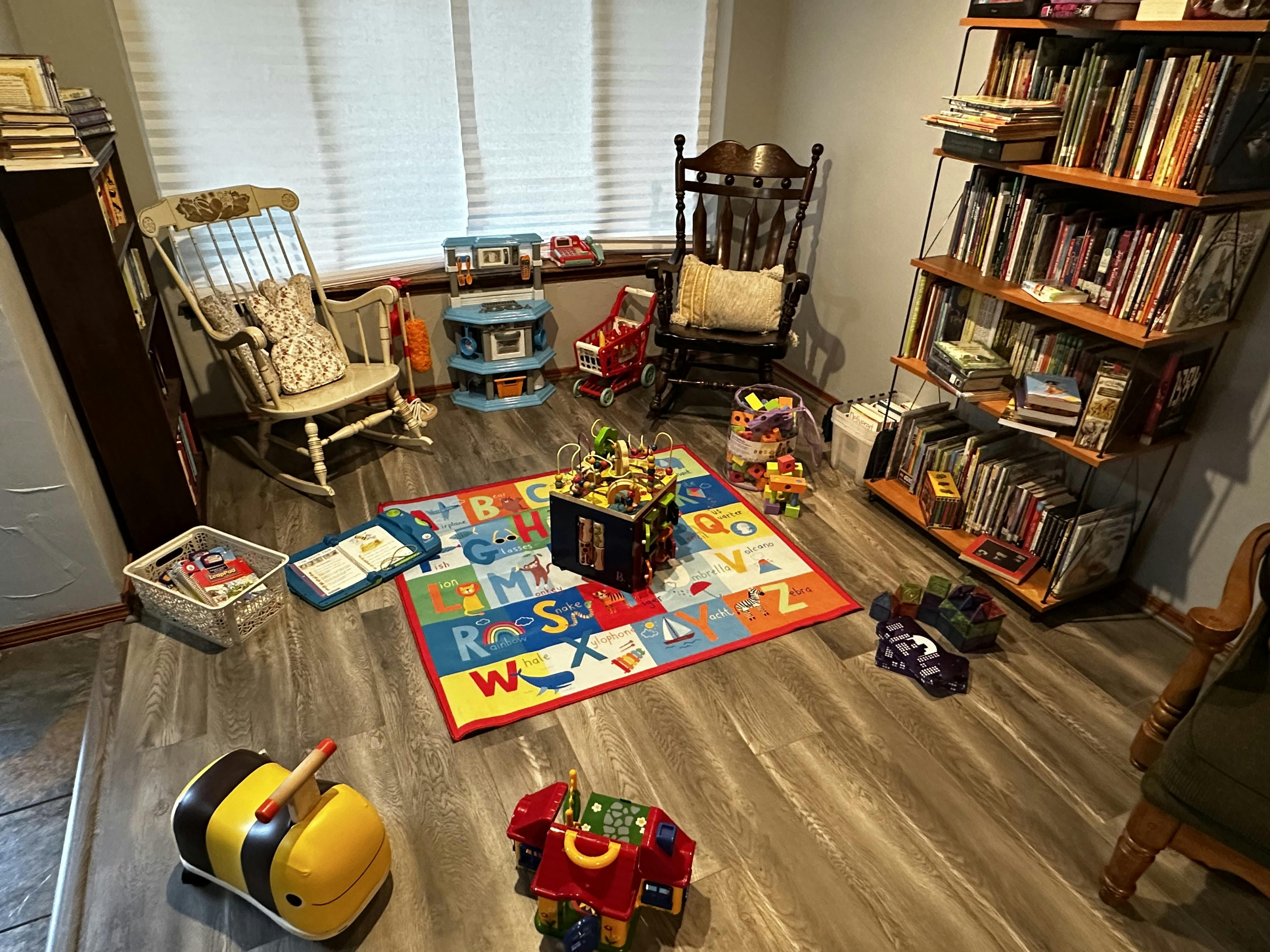 Children's play area with alphabet rug, toys, two rocking chairs, and bookshelves