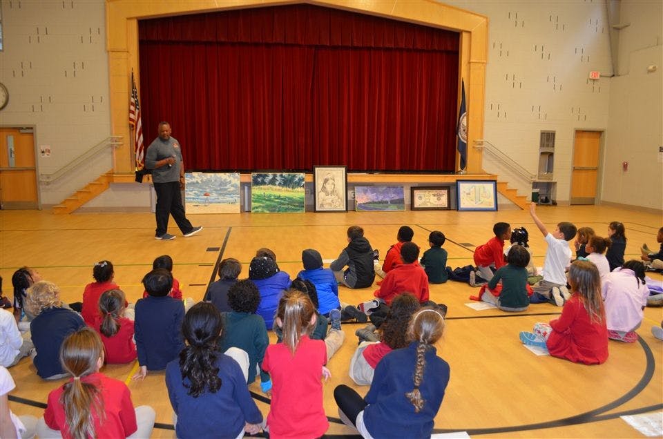 Person speaking to children sitting on a gym floor in front of a stage