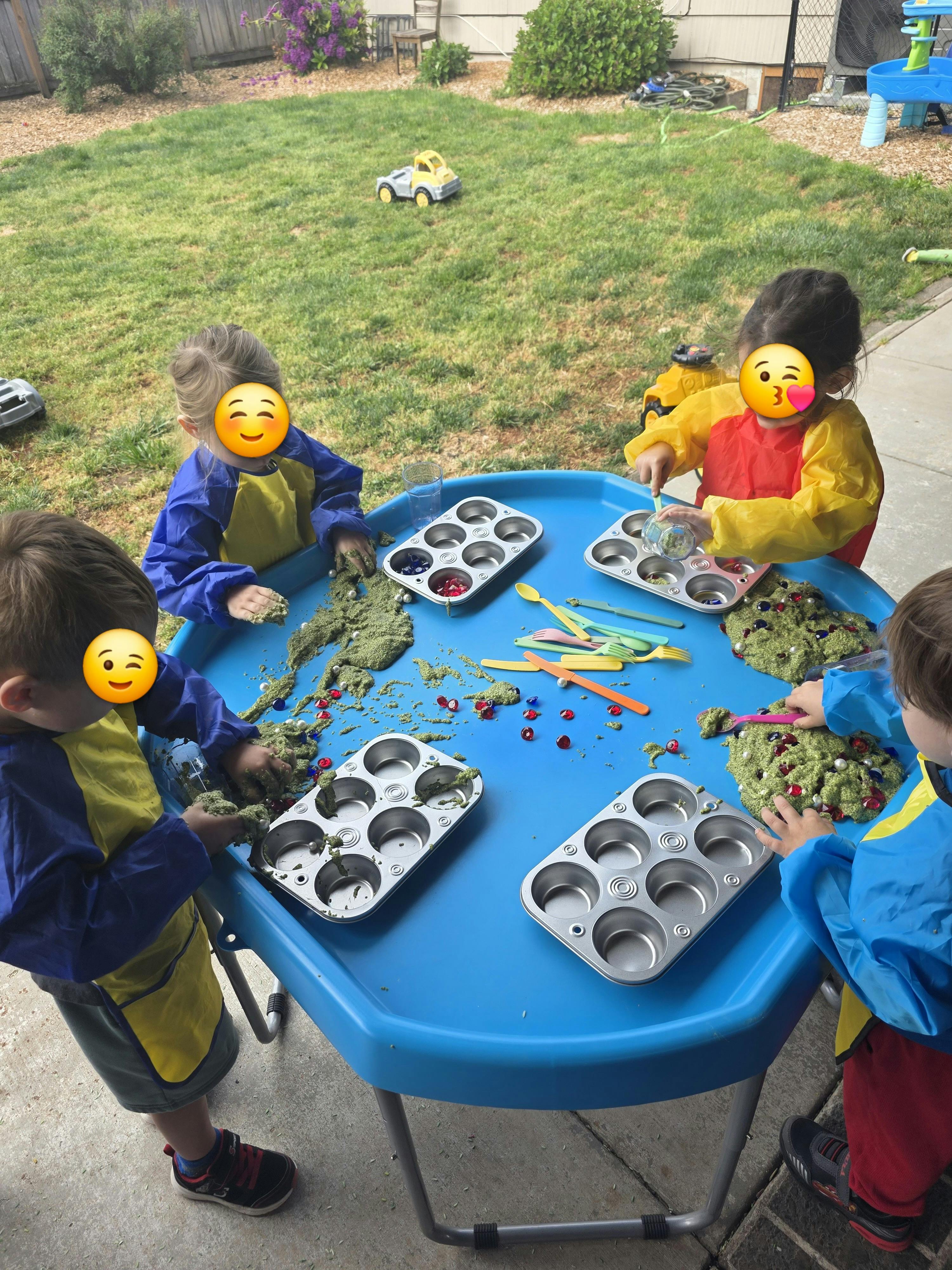 Children playing with green sensory material in muffin tins at a blue table outdoors