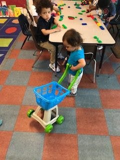 Child pushing a toy shopping cart, another child at a table with blocks