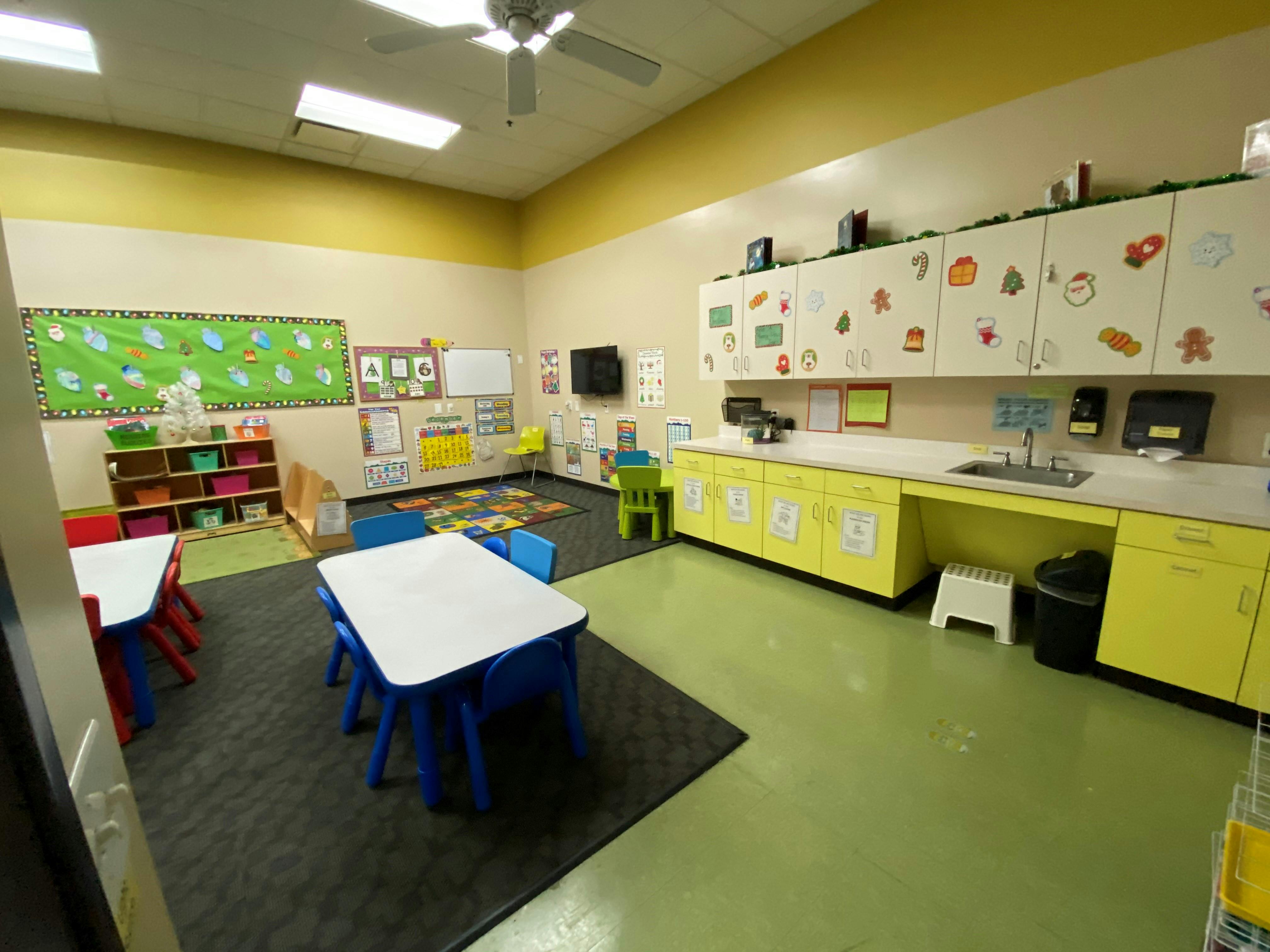 Classroom with small tables and chairs, green bulletin board, and yellow cabinets