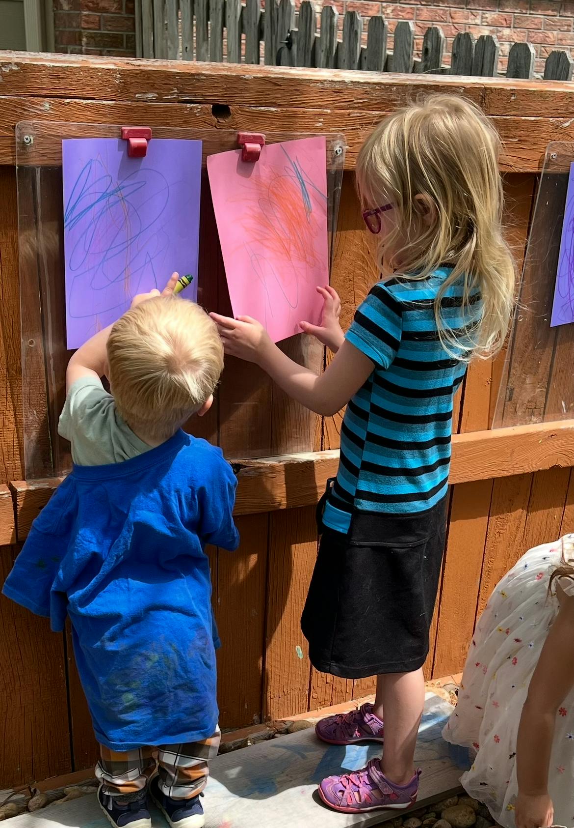 Two children drawing on paper attached to a wooden fence