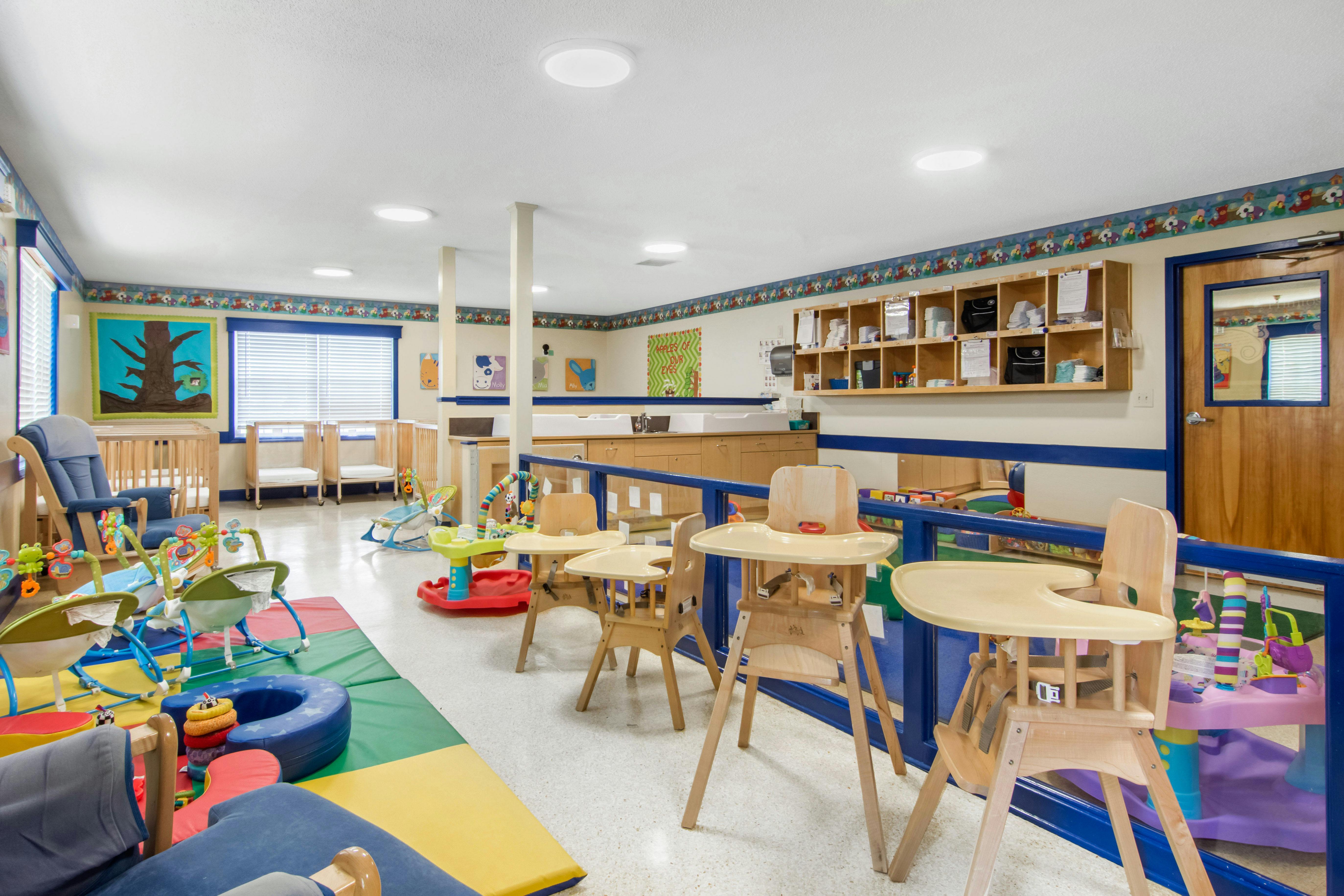 An empty daycare room with high chairs, cribs, and baby toys