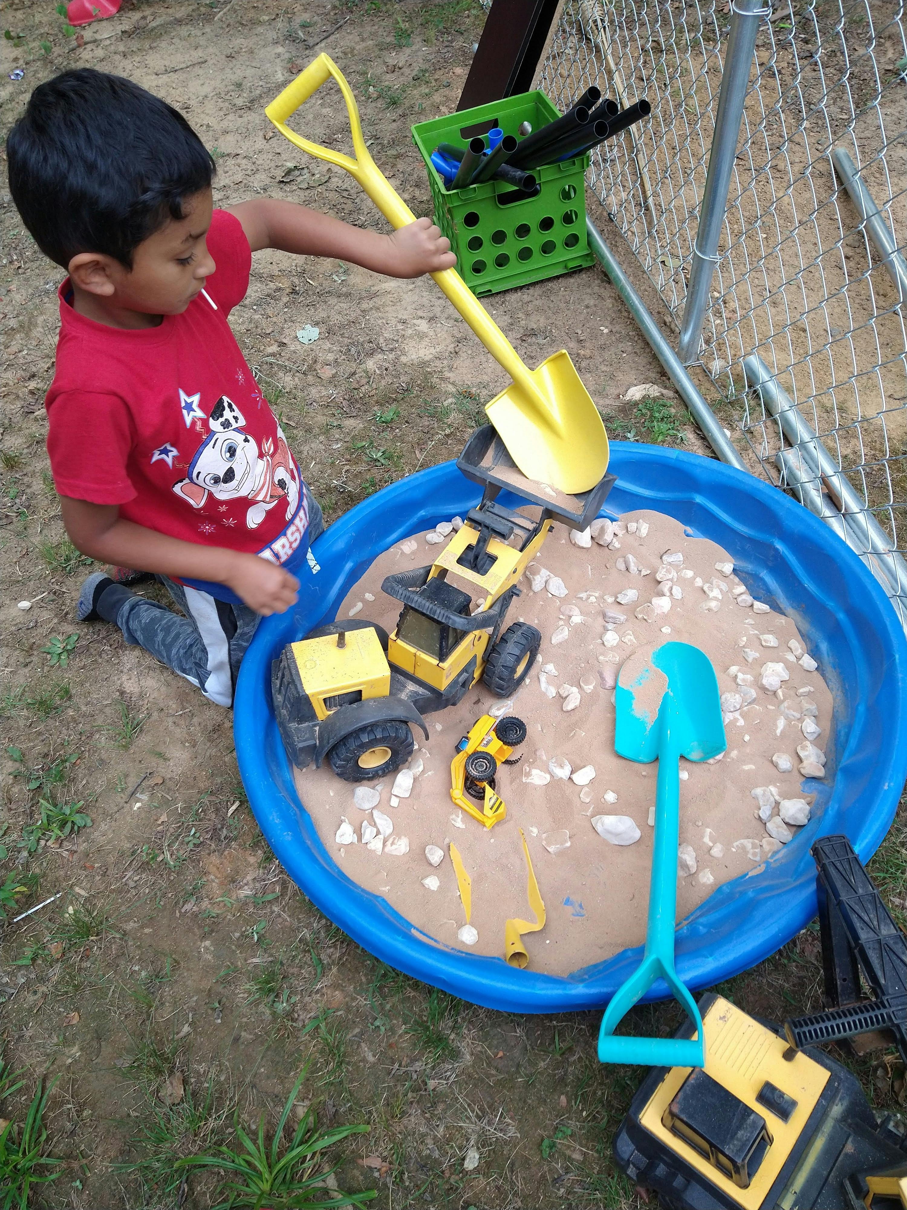 Child playing with a yellow shovel and toy construction vehicles in a sandbox