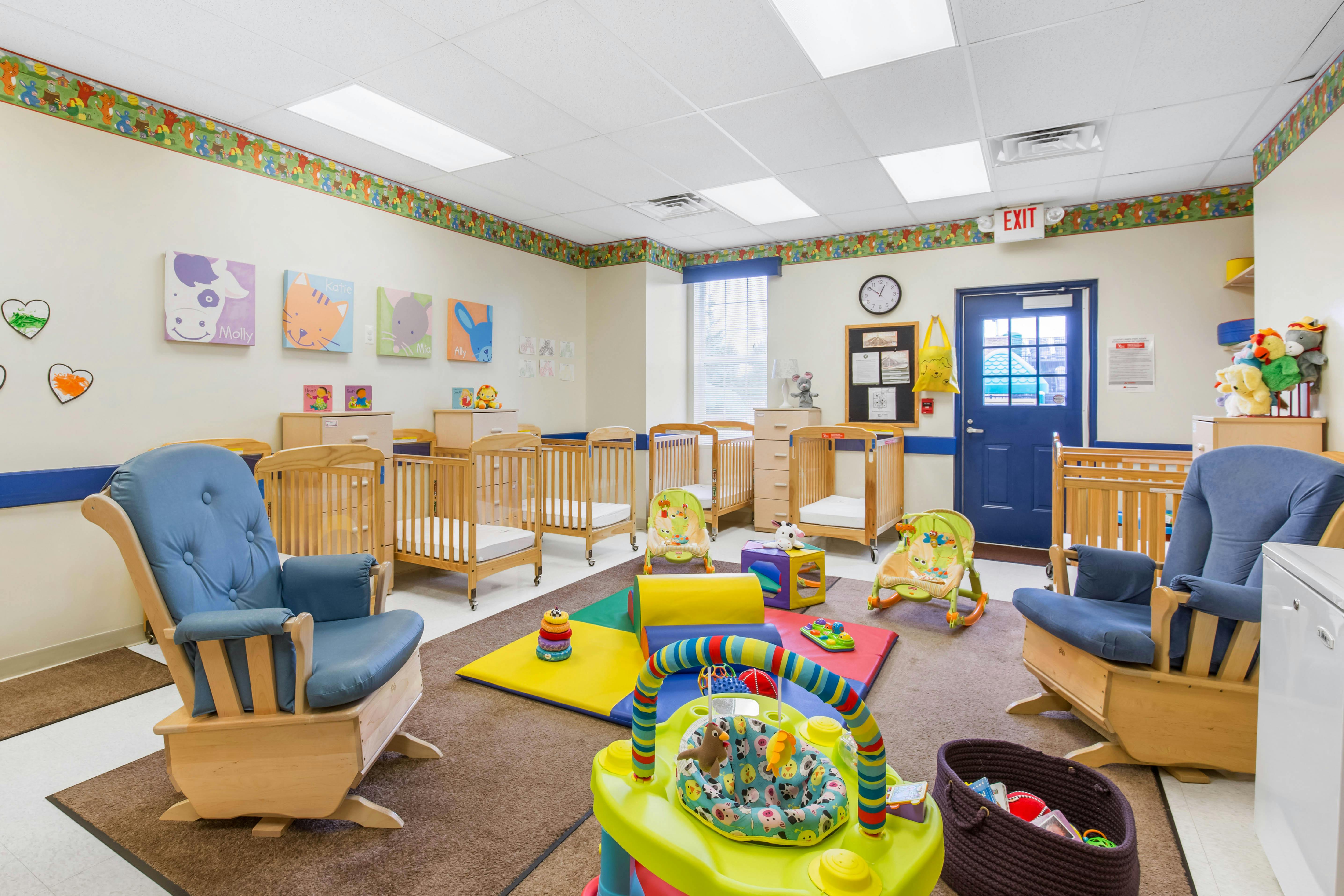 Classroom with multiple cribs, two blue rocking chairs, and colorful play mats and toys