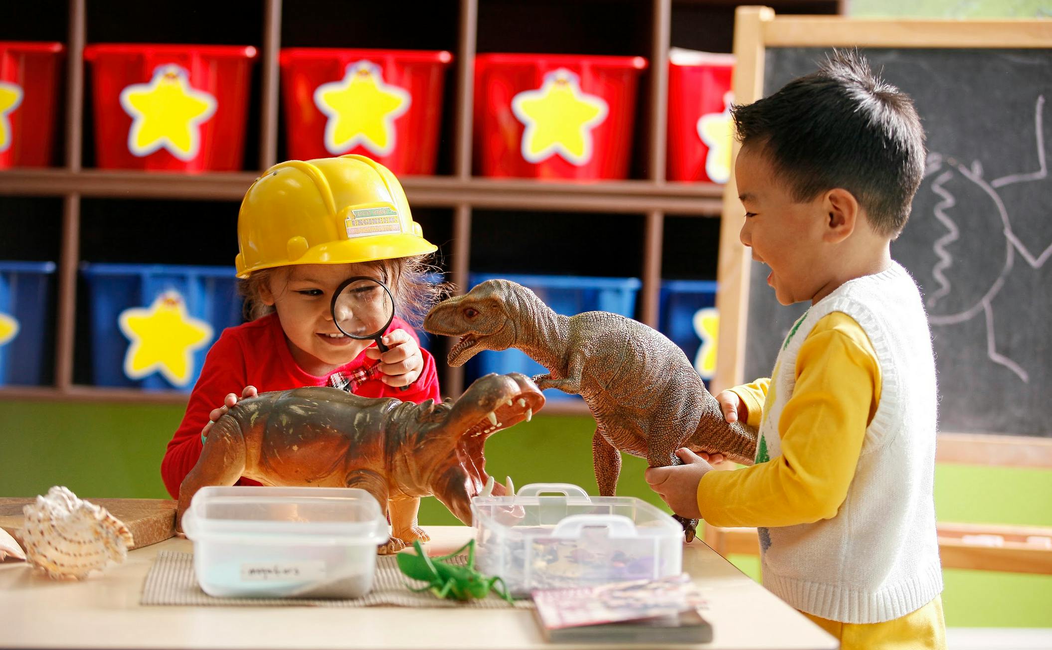 Child in hard hat looking at dinosaur toy with magnifying glass, another child nearby