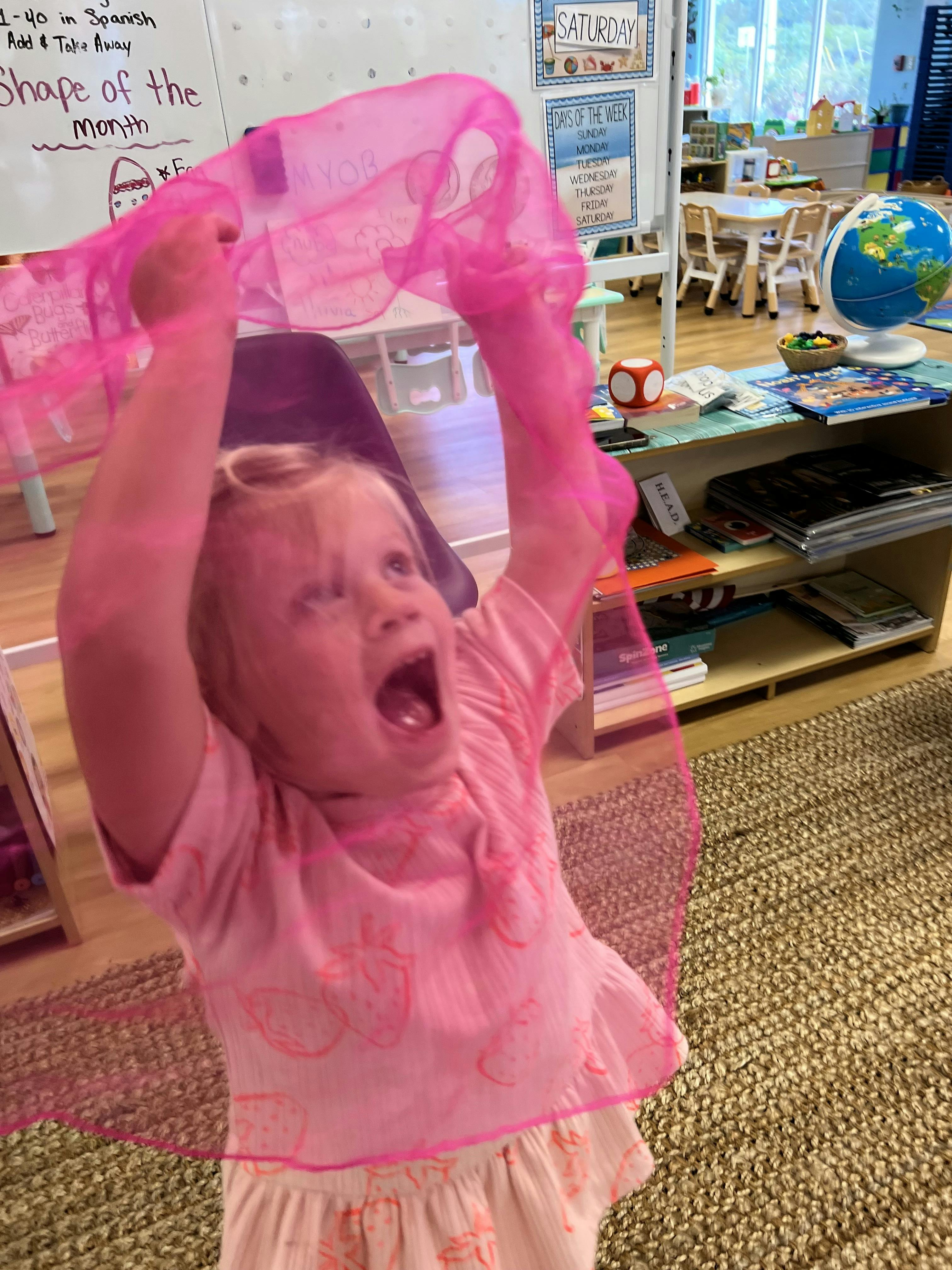 Child in a classroom holding up a pink translucent scarf
