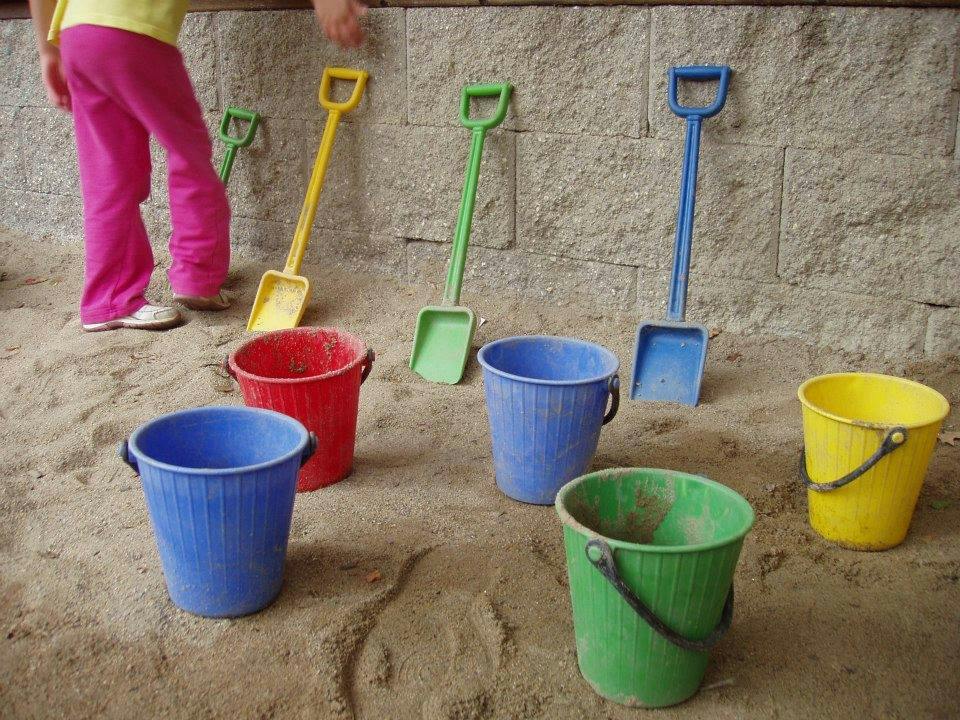 Child standing in a sandbox with colorful buckets and shovels
