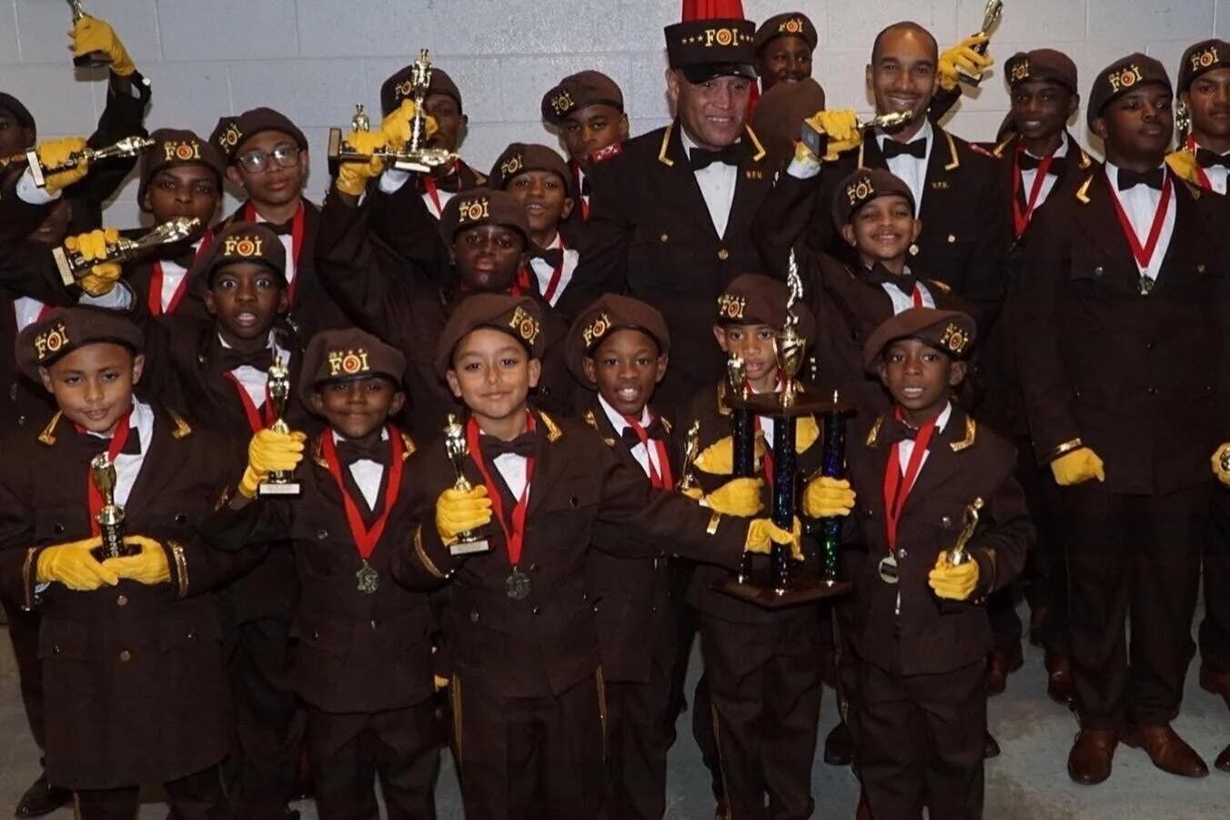Group of children and adults in brown uniforms with hats marked FOI, holding trophies