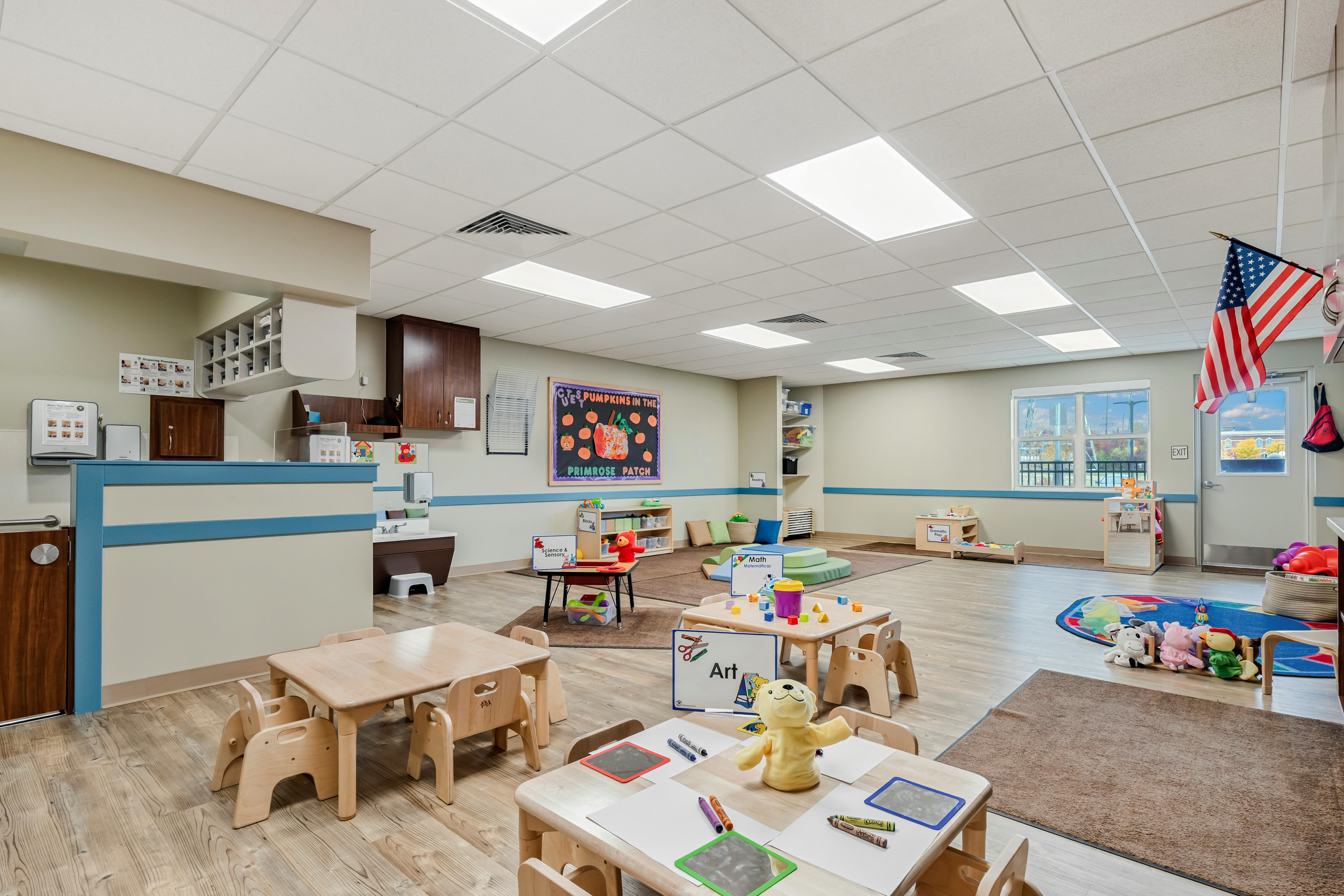 Classroom with small tables and chairs, play mats on the floor, and a colorful bulletin board