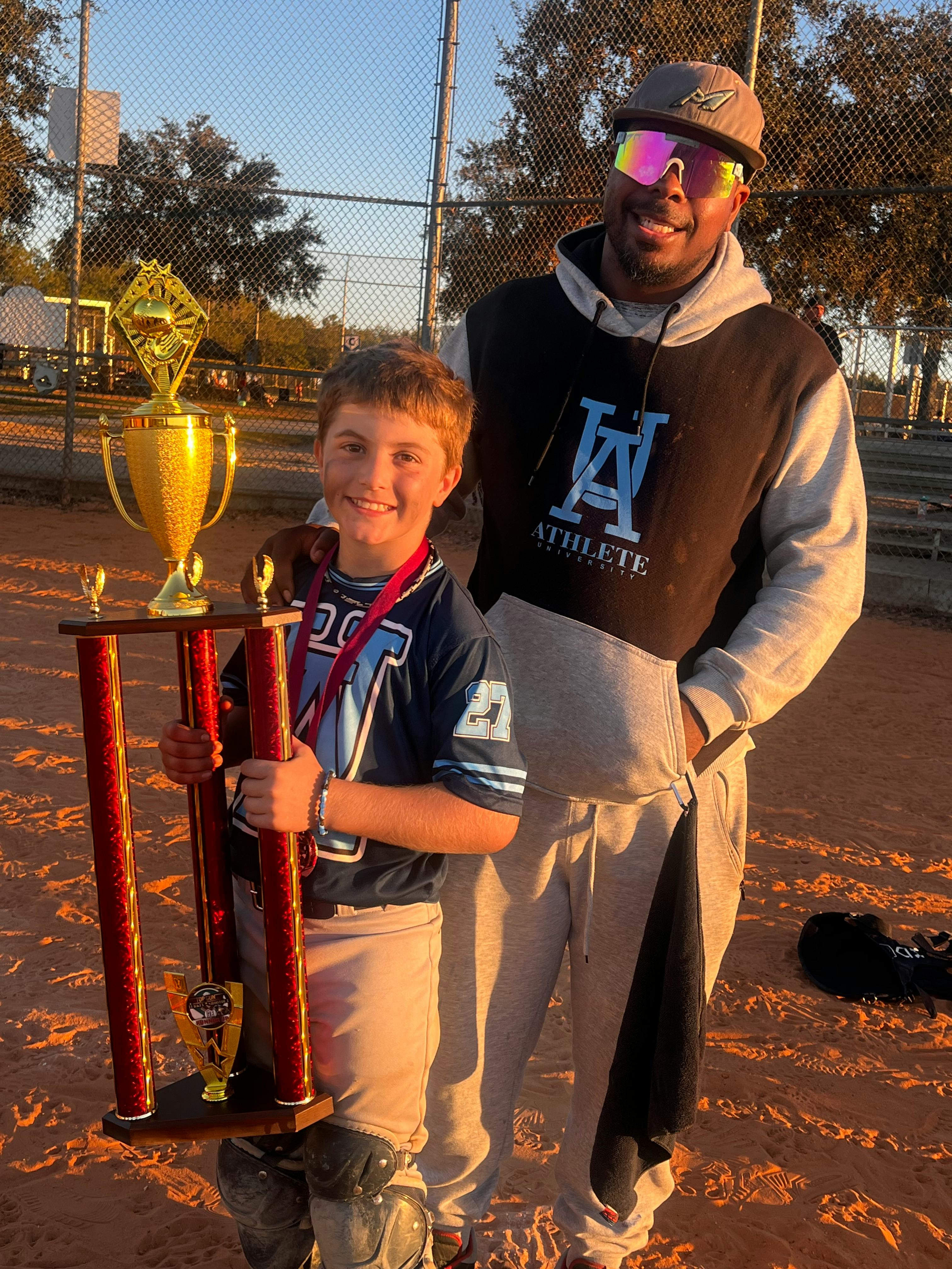 Child in baseball uniform holding a trophy next to an adult