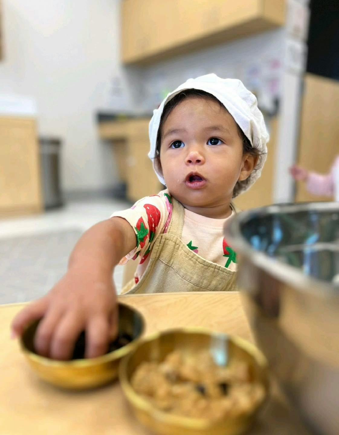 Child in a white cap reaching into a small bowl on a table