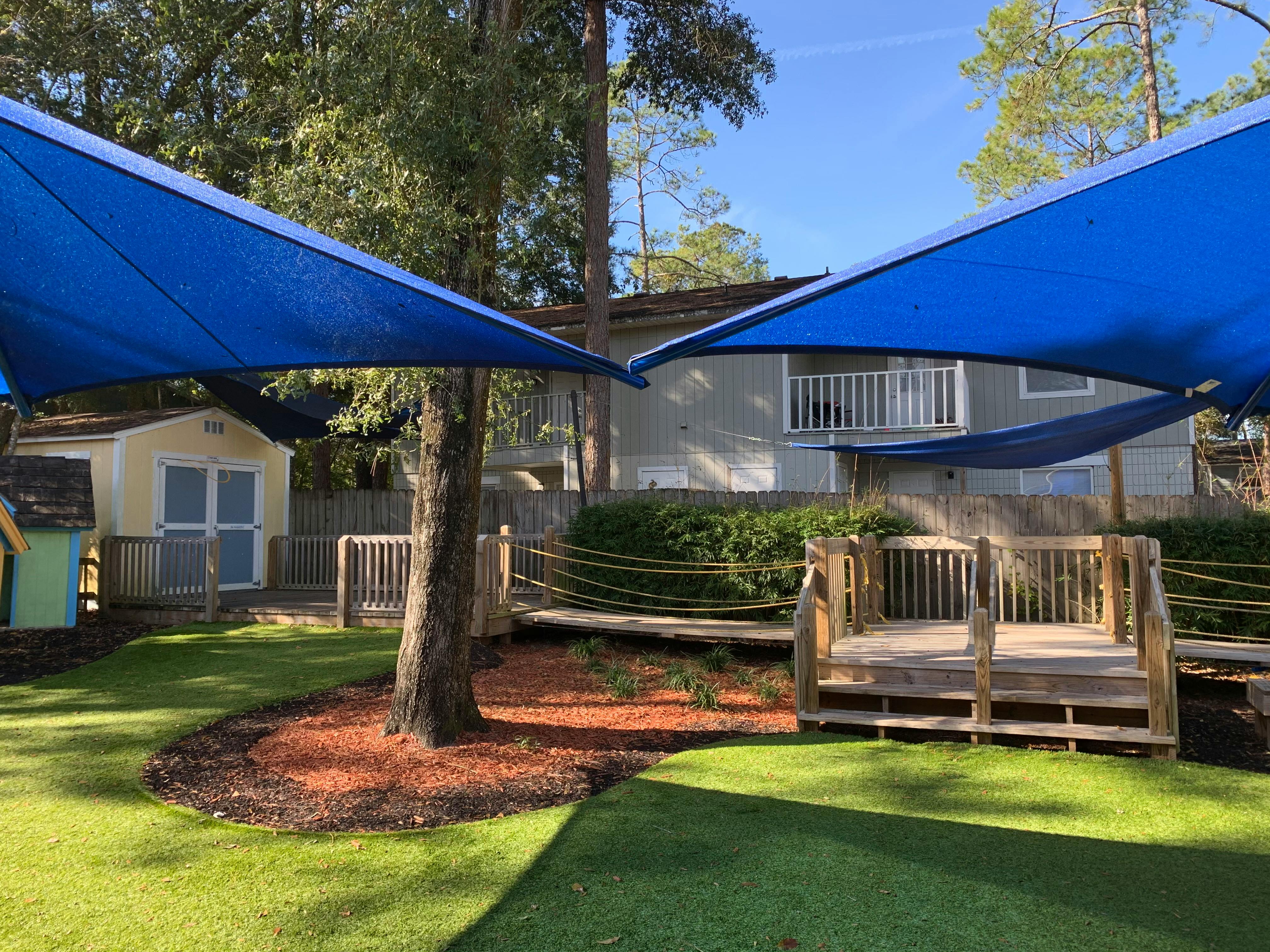 Outdoor play area with blue shade sails, green turf, wooden deck, and shed