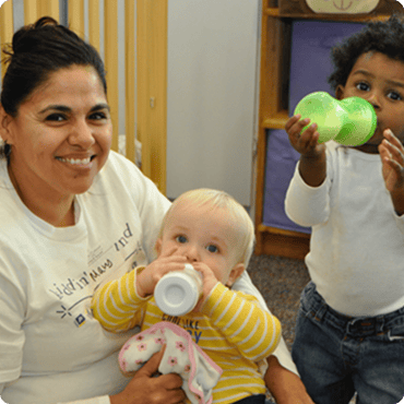 Adult holding a child drinking from a bottle, another child holding a toy