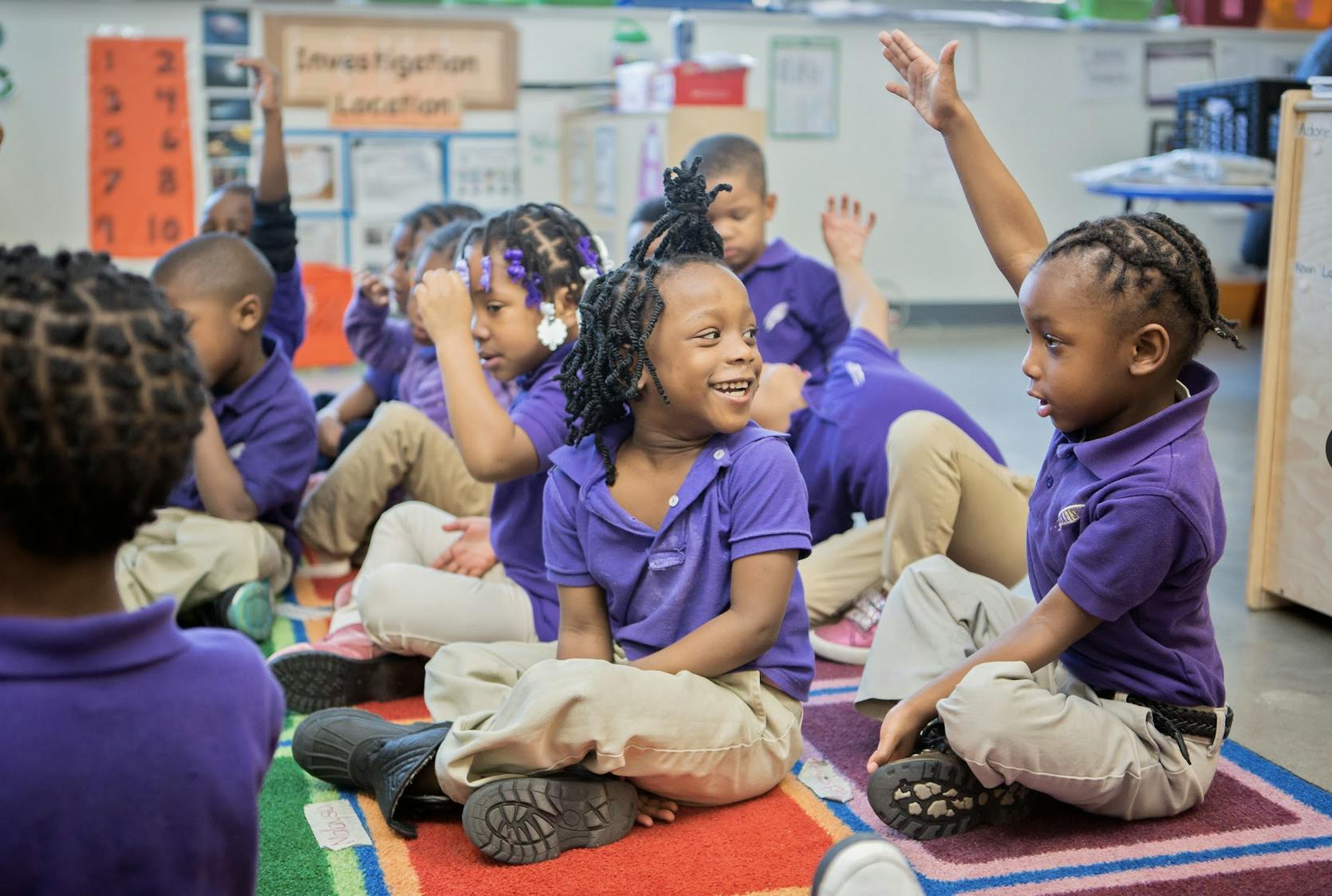 Children sitting on a rug in a classroom, some with hands raised